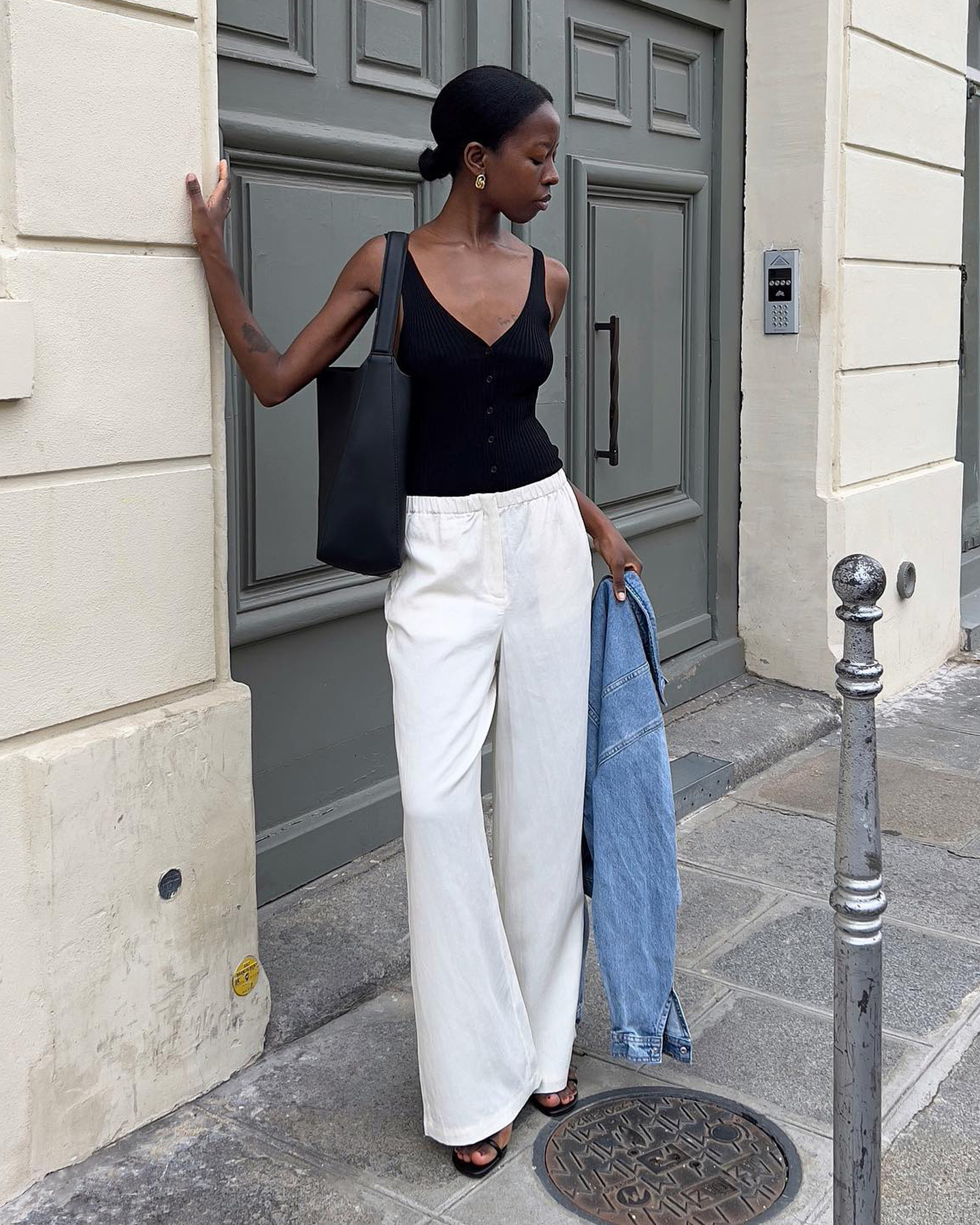 style influencer Sylvie Mus leans near an ornate door on a street in Paris, France wearing a low-cut black tank top, black shoulder tote bag, white linen drawstring pants, and strappy sandals with a denim jacket in her hand