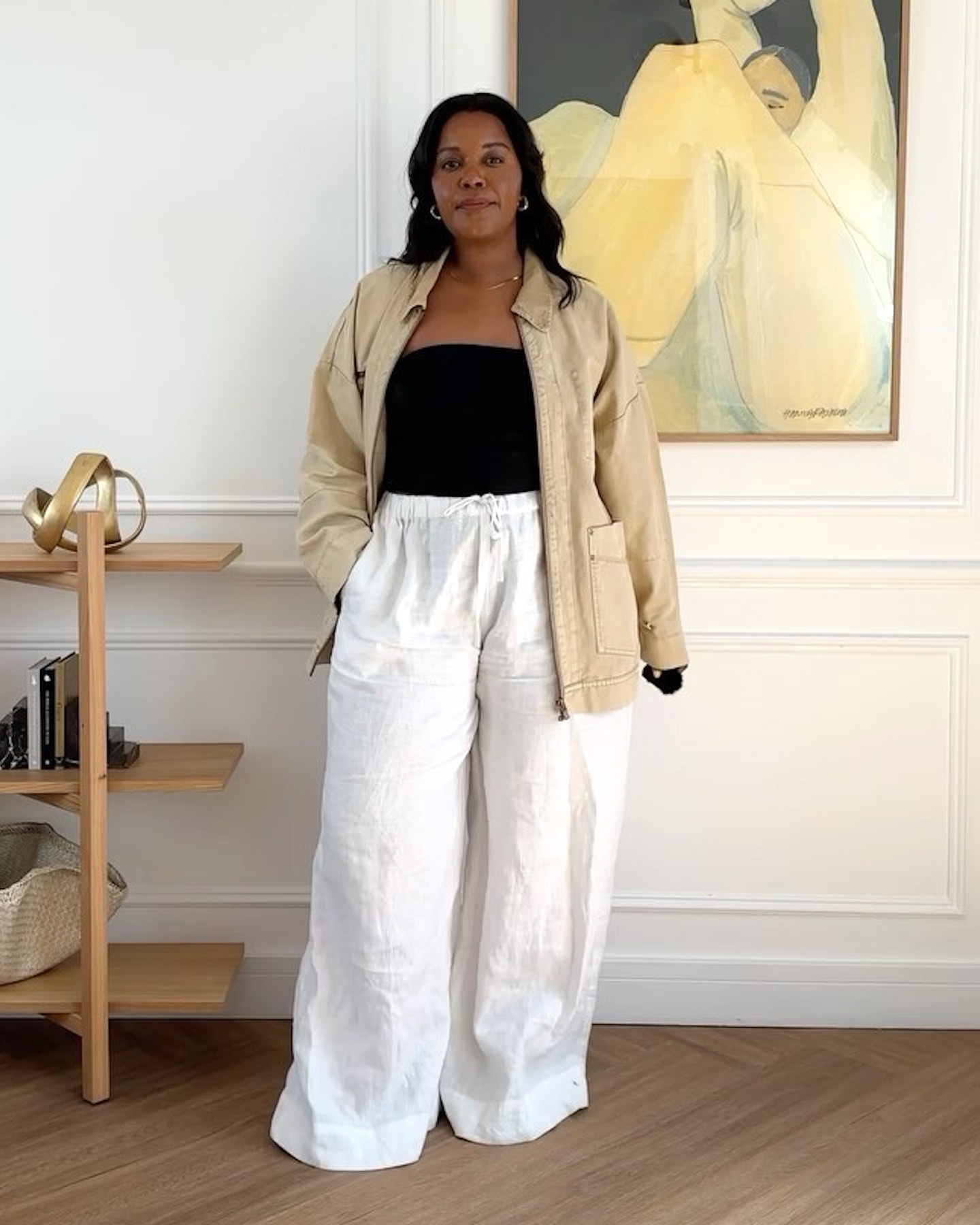 British style influencer Karina Marriott poses in her chic apartment in front of abstract art wearing a tan barn coat, black basic top, and white wide-leg linen pants.