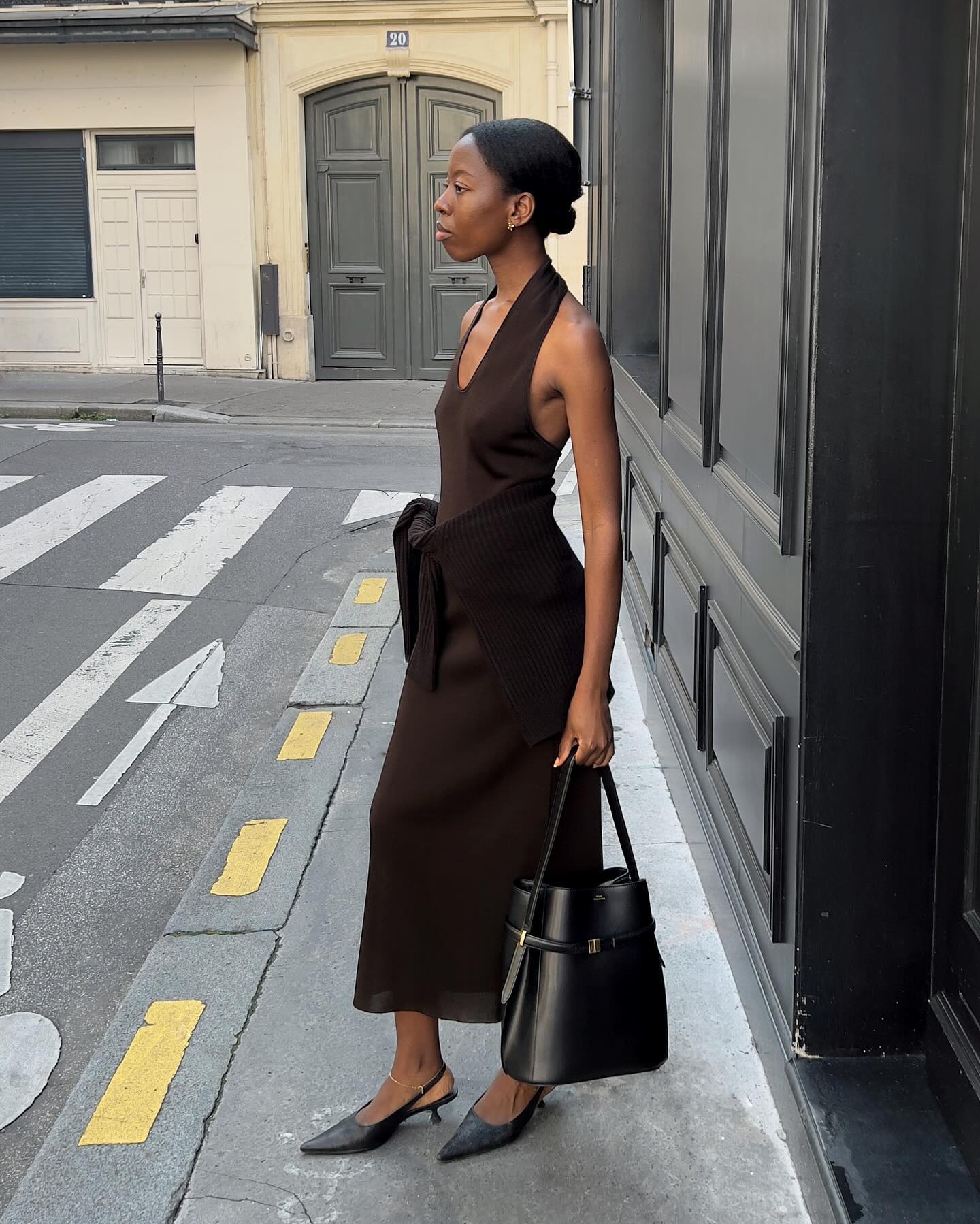 European style influencer poses on a Paris sidewalk wearing a brown halter dress with a brown sweater tied around her waist holding a black leather Toteme tote bag and black kitten-heel slingbacks.