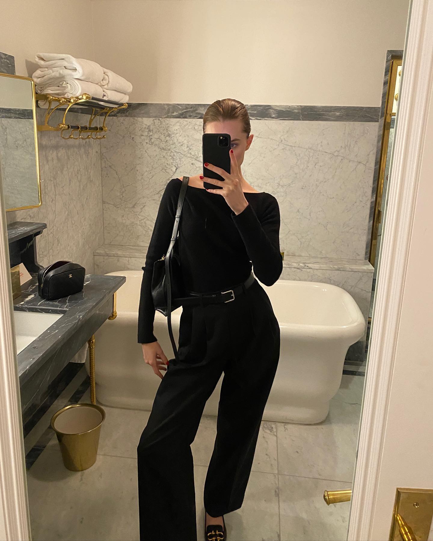 NYC style influencer Christie Tyler poses for a mirror selfie in a marble bathroom wearing a black boatneck top, black belt, black pants, loafers, and a leather shoulder bag.