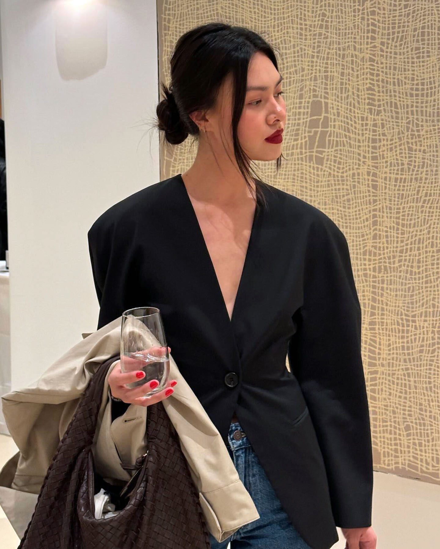 Style influencer Mimi Nguyen poses with a low bun, red lipstick, black collarless cinched-waist blazer, and jeans with a trench coat and brown woven bag on her arm.
