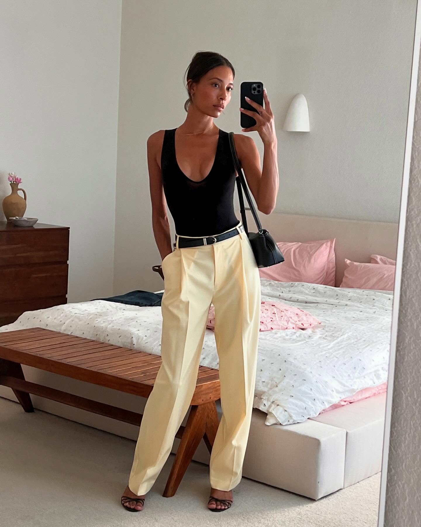 fashion influencer Tylynn Nguyen poses for a mirror selfie in her bedroom wearing a low-cut black top, black belt with silver hardware, a black east-west bag, butter-yellow pants, and black strappy sandals
