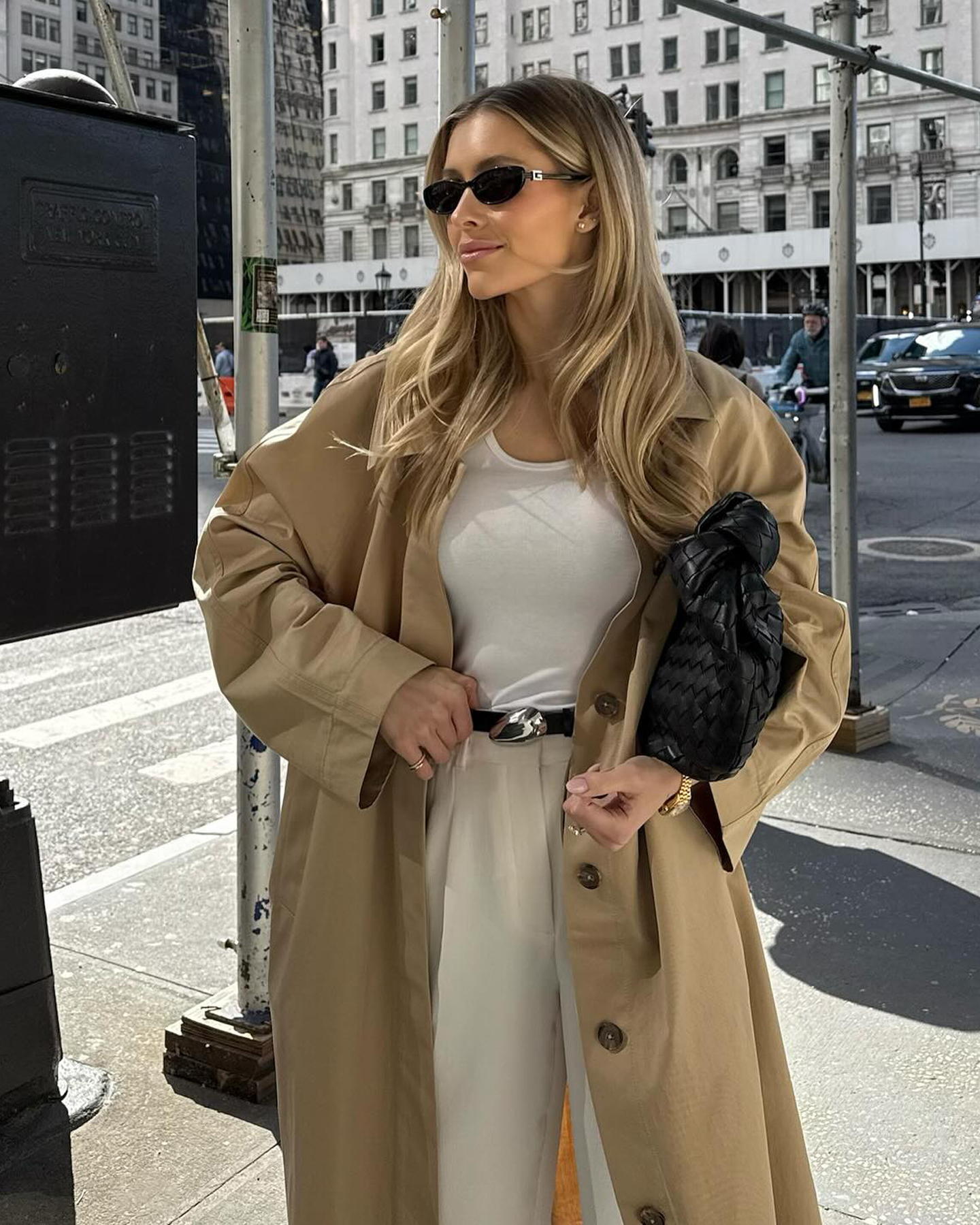 fashion influencer Fredricka Ekerot poses on a New York City sidewalk wearing black oval sunglasses, a black leather woven bag, white basic top, black belt with silver sculptural belt buckle, beige linen pants, and an oversize trench coat
