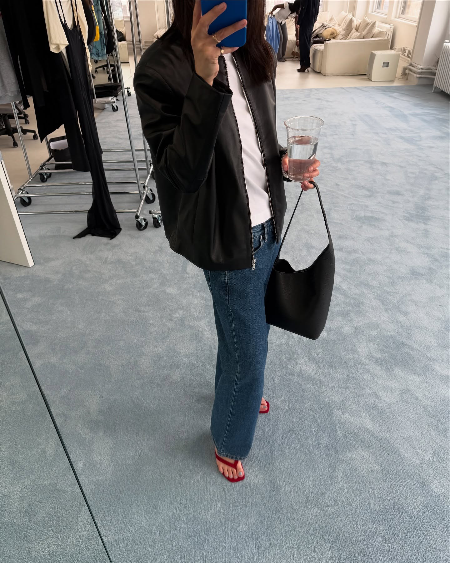 fashion influencer Mimi Nguyen poses for a mirror selfie in a store with light blue carpets wearing a black leather jacket, white T-shirt, simple black leather tote bag, straight-leg jeans, and red heeled flip-flop sandals