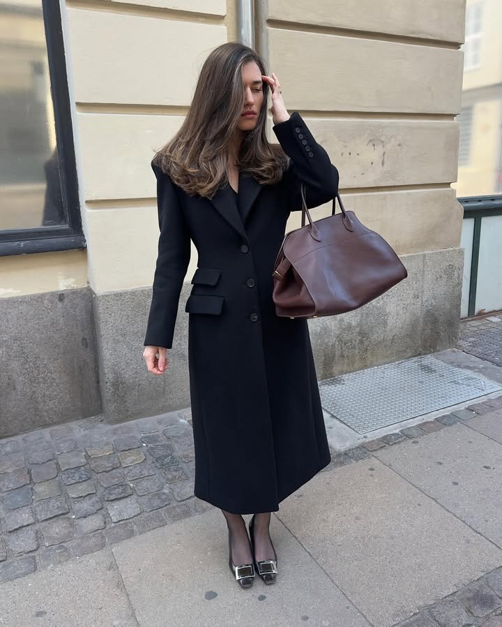 Woman standing and fixing her hair, wearing a black trench and carrying a large brown purse.