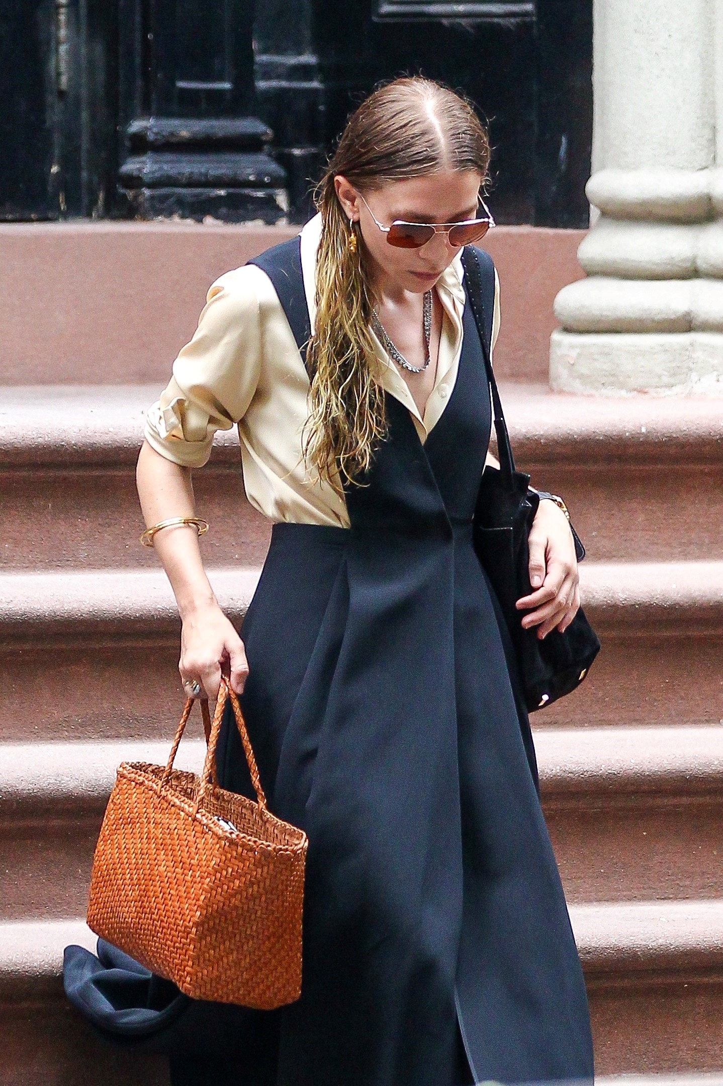 Ashley Olsen spotted walking down brownstone stairs wearing aviator sunglasses, a tan blouse layered under a navy blue sleeveless dress, a black patent shoulder bag, and camel brown Dragon Diffusion woven tote bag