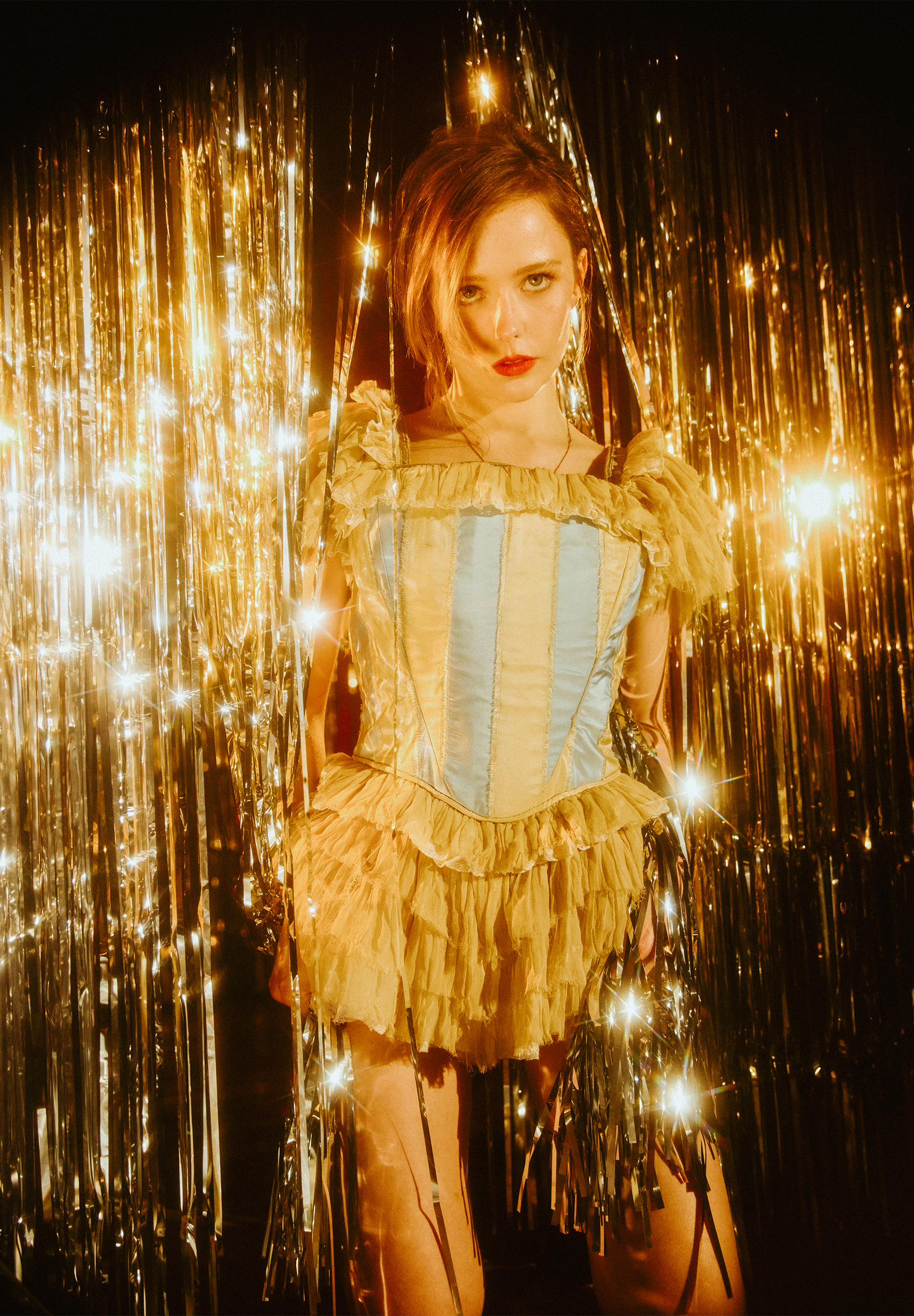 Suzanna Son from Fear Street: Prom Queen is standing in a silver tinsel backdrop wearing a Gabe Gordon minidress with ruffled skirt and gold and blue striped bustier top.