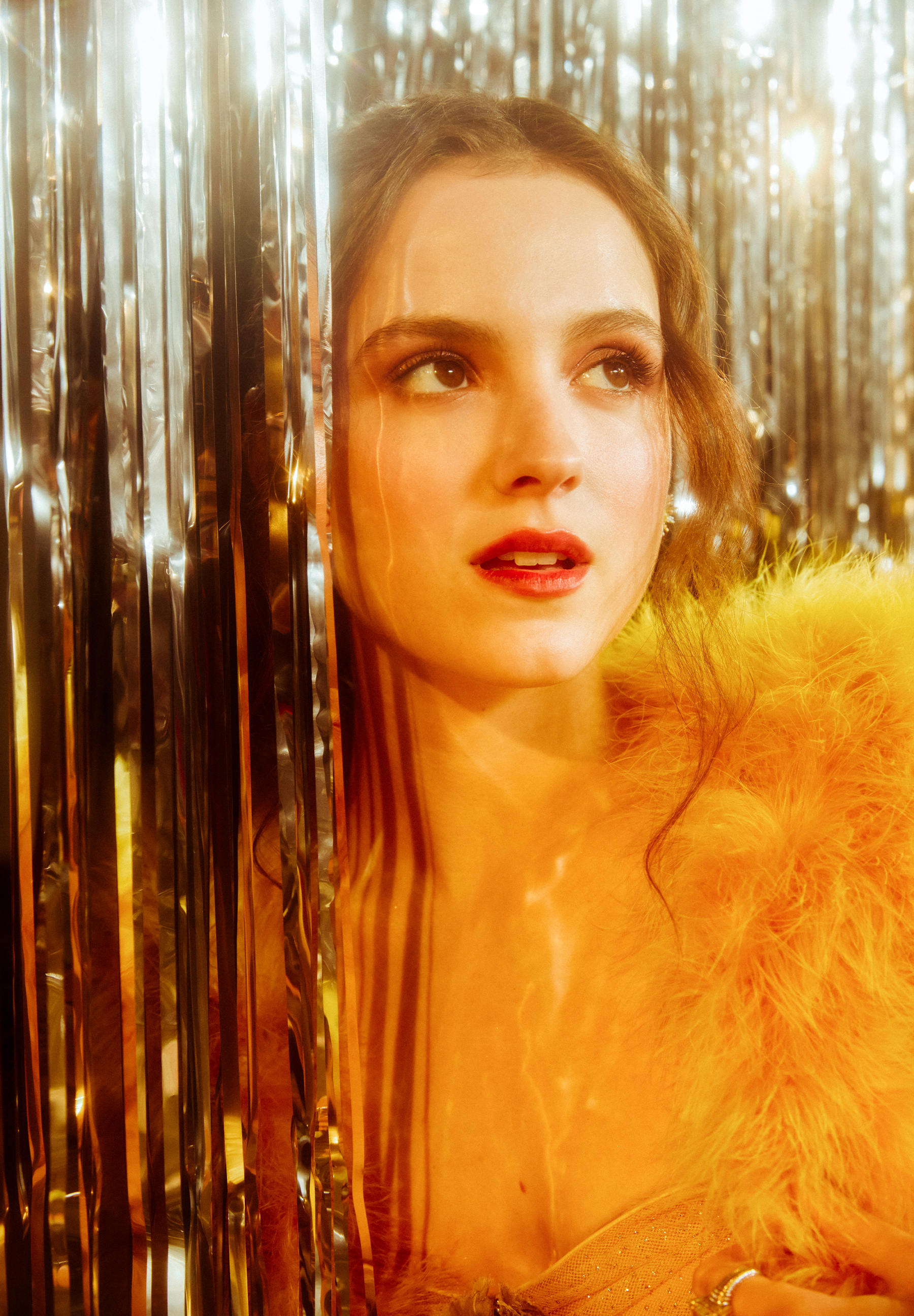 Fear Street: Prom Queen actress Fina Strazza poses with a silver tinsel backdrop wearing a golden yellow Gucci gown with furry trim.