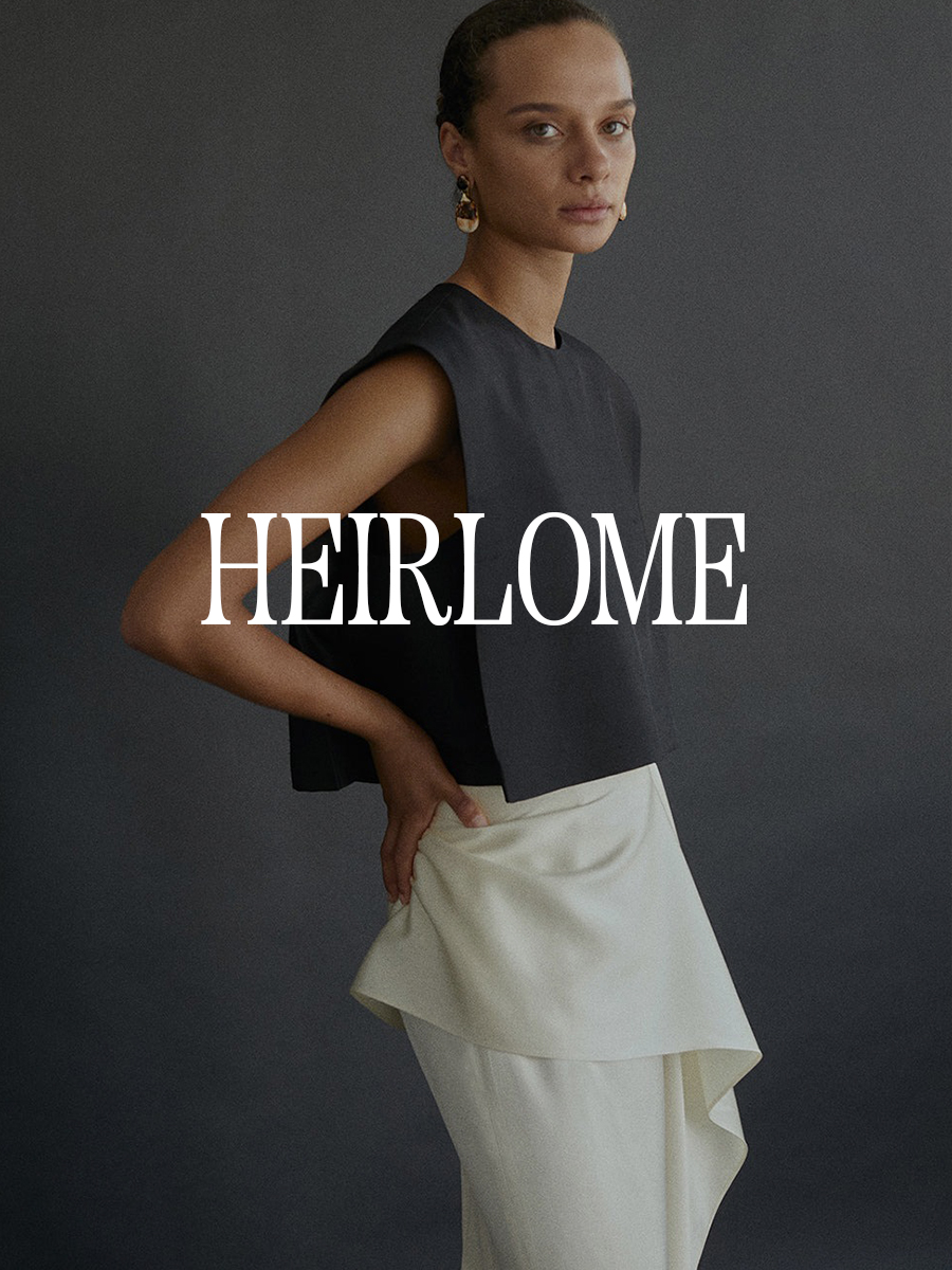 A photo of a model wearing a gray sleeveless top and cream skirt. Text says "Heirlome" in white capital serif letters.