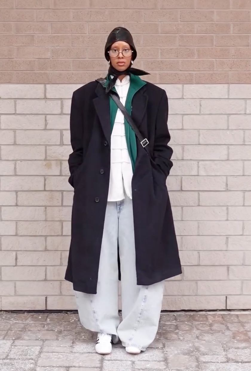 A woman standing against a brick wall and wearing a leather cap with a long black coat, green sweatshirt, a white button-down, baggy barrel leg jeans, and white sneakers.