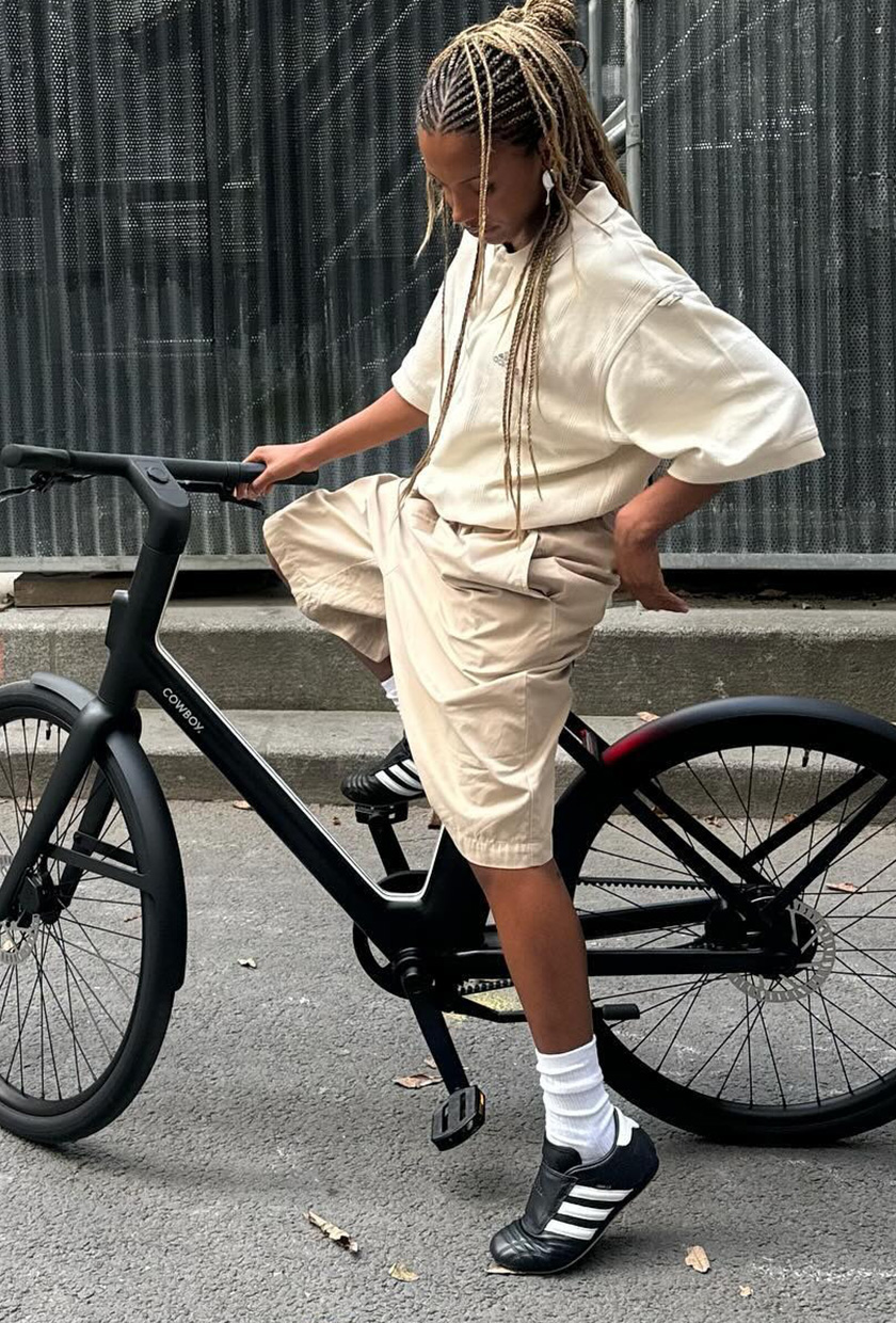A woman sitting a black bike and wearing an oversize tan t-shirt with baggy cargo shorts, white socks, and Adidas sneakers.