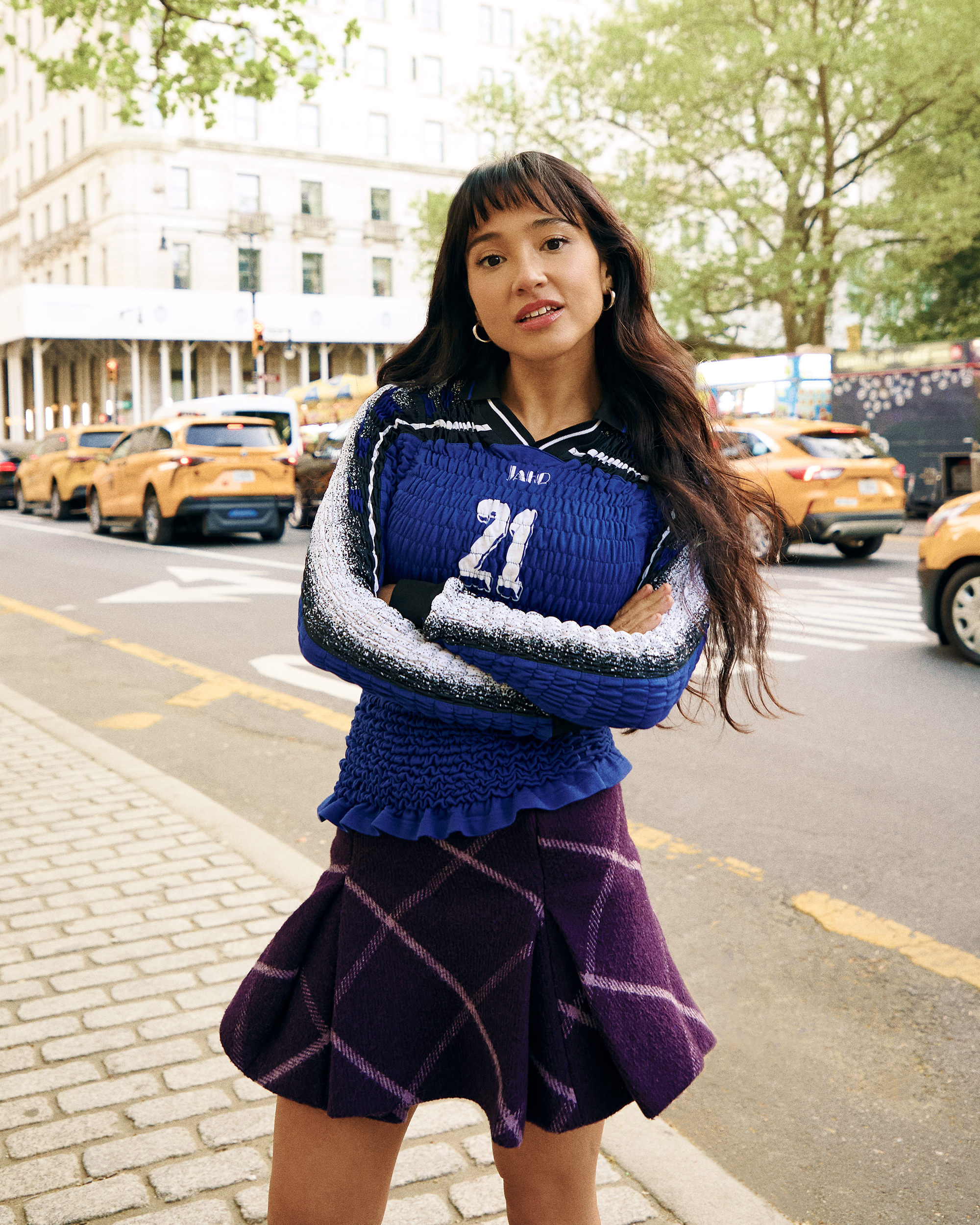Overcompensating actress Wally Baram poses for portrait at the Plaza Hotel in NYC. She is wearing a long-sleeve smocked blue shirt with the number 21 on the front and a purple plaid miniskirt with black leather boots that hit just below the knee.