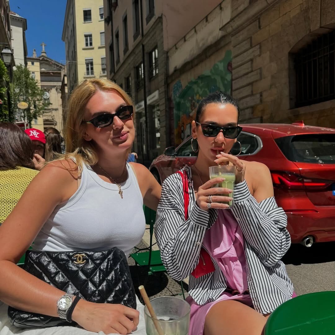 Dua Lipa wears the light pink dress trend in a ruffled dress form. She pairs her dress with a black and white stripped linen shirt a red shoulder bag, black cat eye sunglasses and silver thin hops. In the image she's sat with a friend and drinking a green matcha. This image was taken from her Instagram recently.