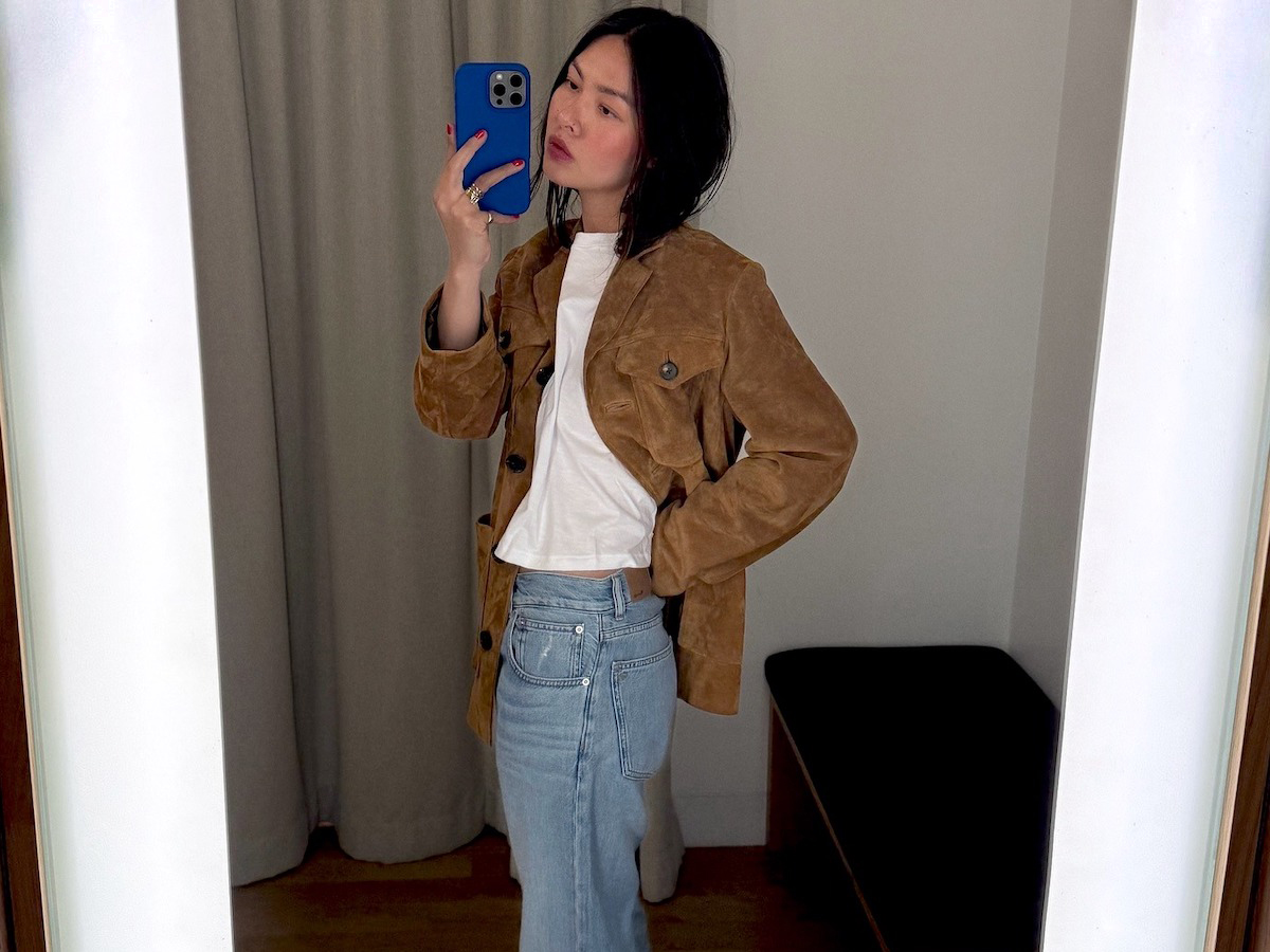 NYC influencer Mimi Nguyen poses for a mirror fitting room selfie in a Madewell store with her bright blue phone case, a brown suede jacket, white tee, and jeans.