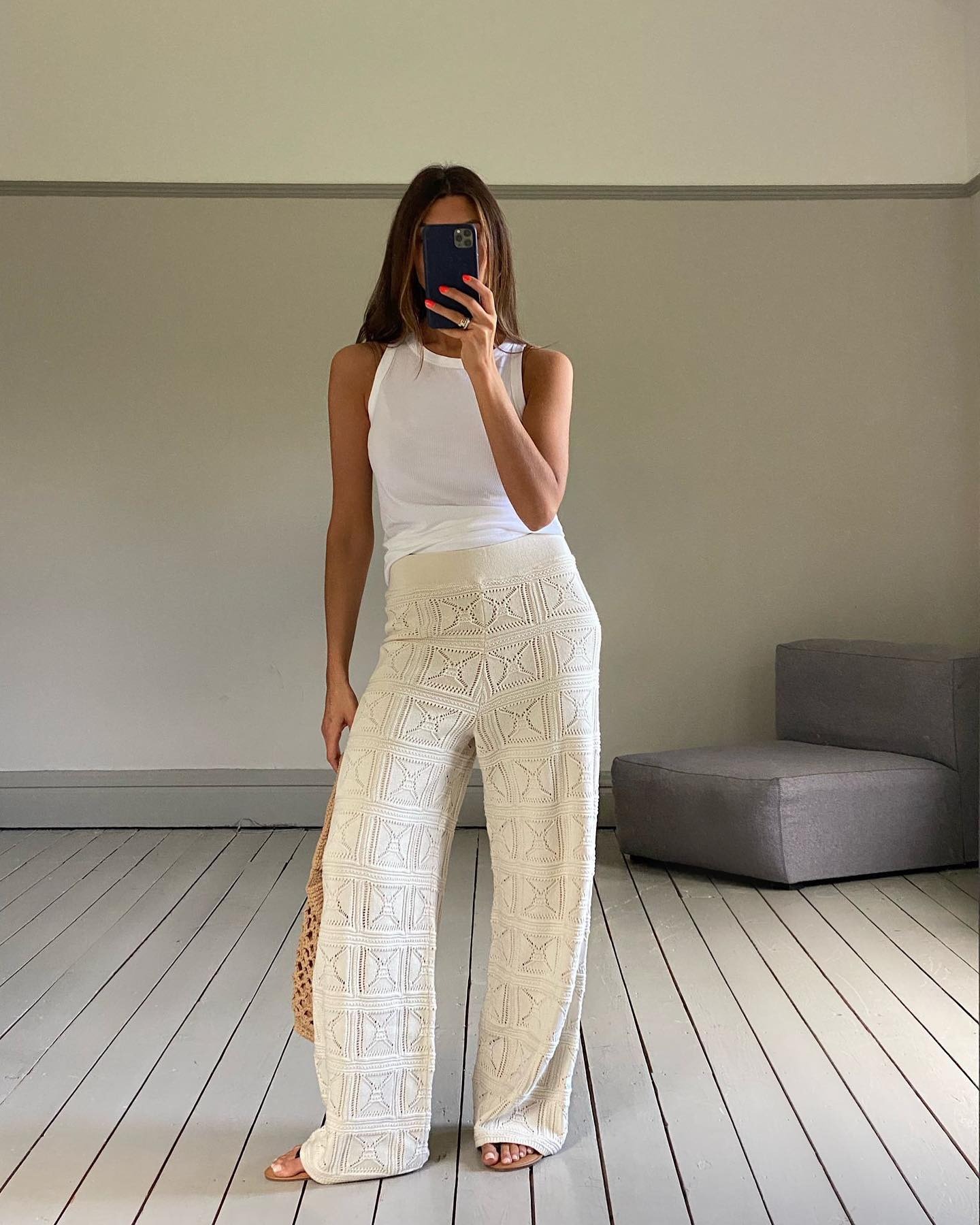 British influencer Marianne Smyth poses for a mirror selfie wearing a white tank top, ivory crochet wide-leg pants, a woven tote bag, and flat sandals.