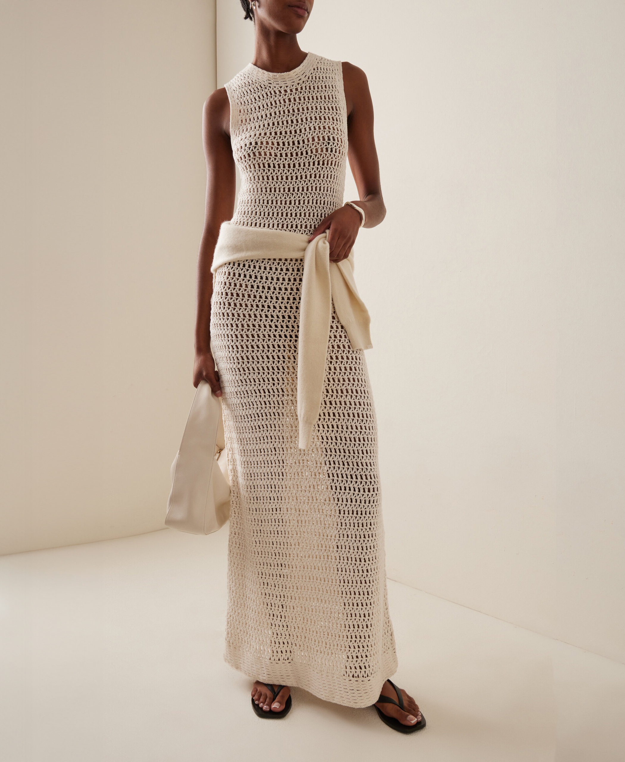Moda Operandi model wearing Leset Lucy crocheted-cotton maxi dress, a sweater tied around the waist, a white mini bag, and flat black flip-flop sandals.