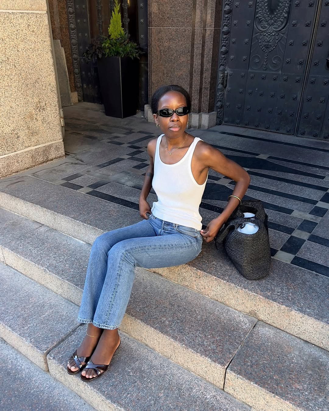 A woman sitting on a stoop wearing a pair of light wash blue jeans, a white tank top, and black kitten heels.