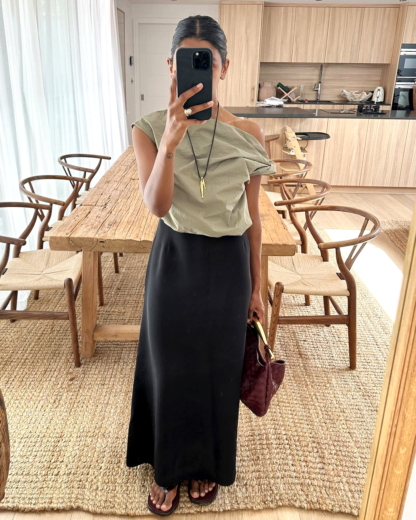 British style influencer Monikh Dale poses for a mirror selfie in her dining room in London wearing a draped off-the-shoulder neutral top, AlighieriBlack & Gold 'The Gone Fishing' Necklace, Bottega Veneta sardine bag, black maxi skirt, and black flip-flop sandals