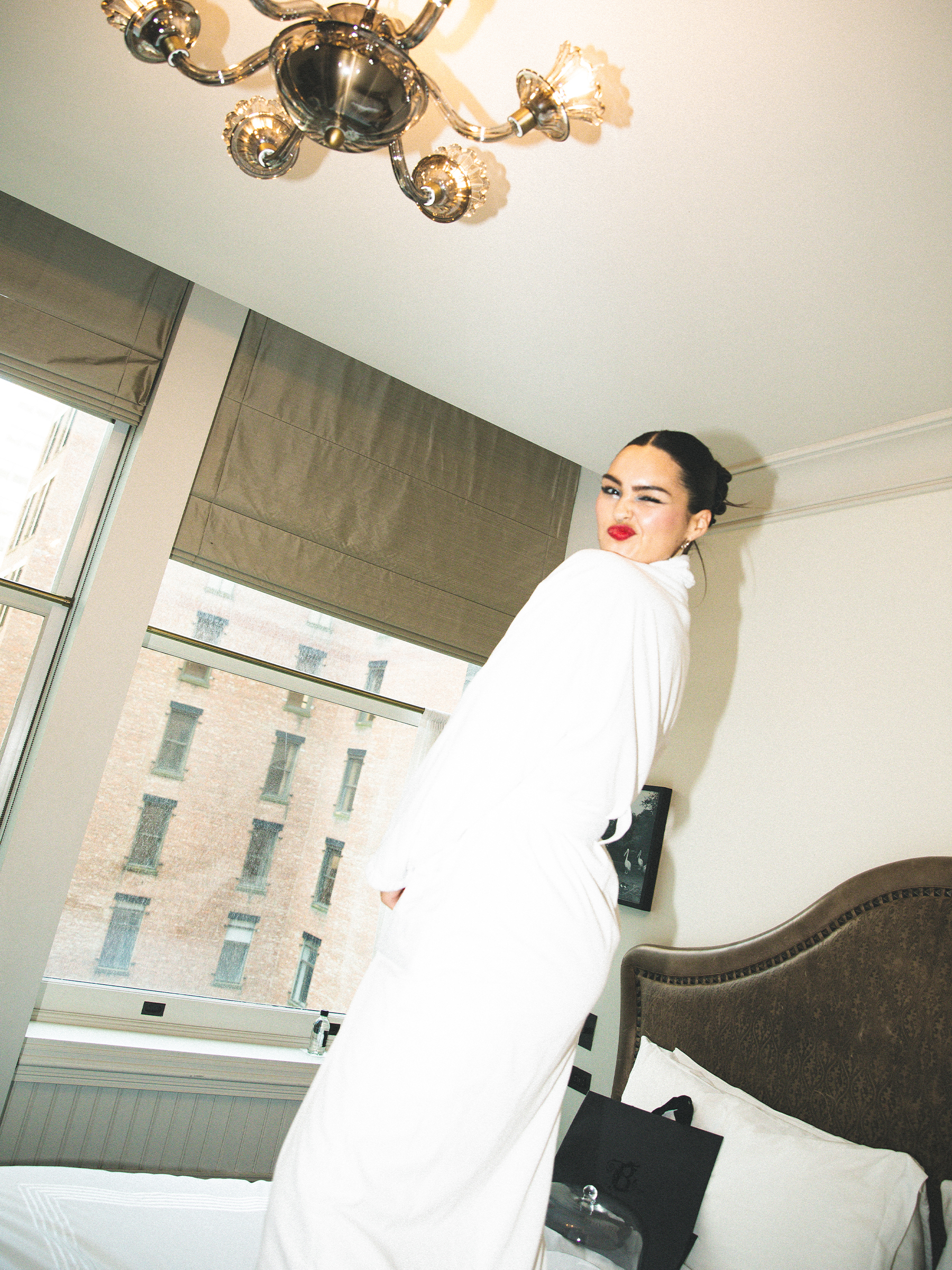Anna Lambe poses for a photo in her hotel room. She is standing on the bed wearing a white robe and is winking at the camera.