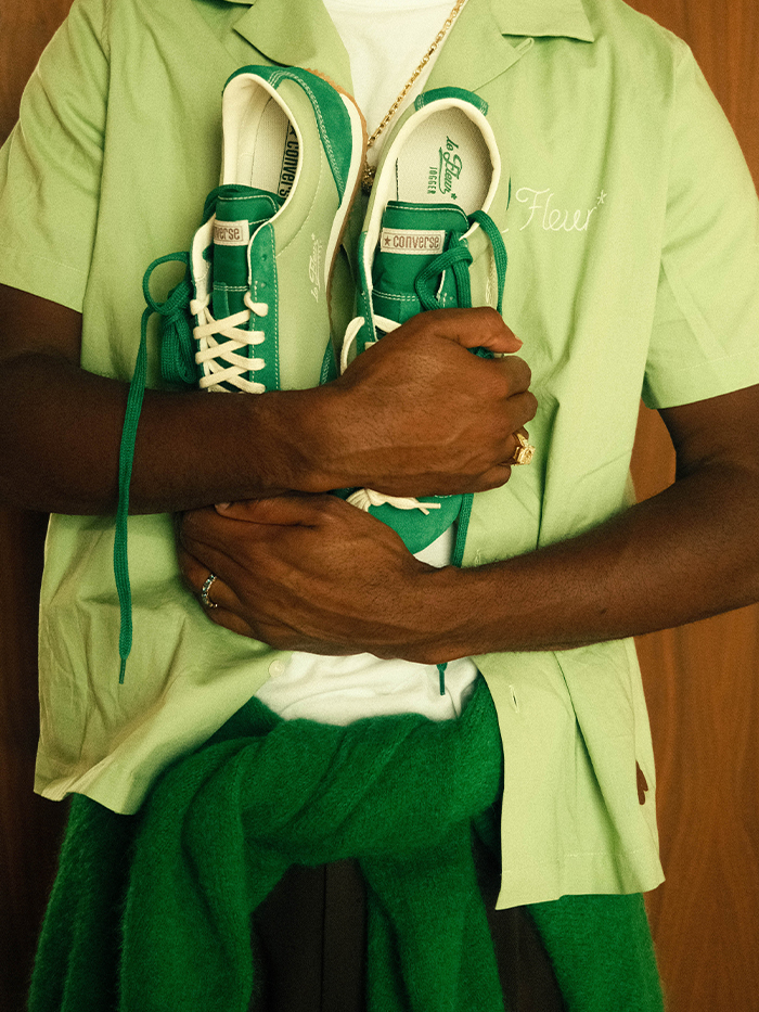tyler hold a green paire of the jogger shoe while wearing a green button down and white t-shirt with a green sweater tied around his waist