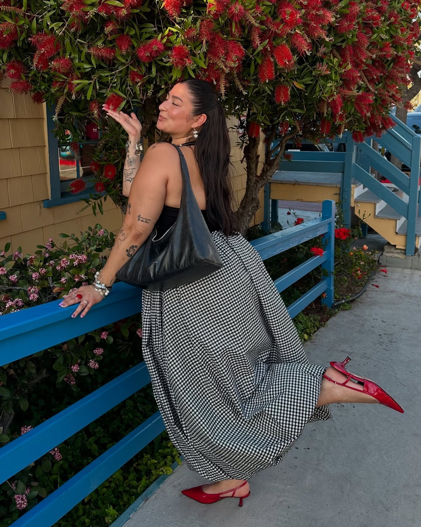 Influencer wears red kitten heels with a gingham print maxi skirt and black top. She is standing outside in front of a flowing tree with a black bag slung over her shoulders.