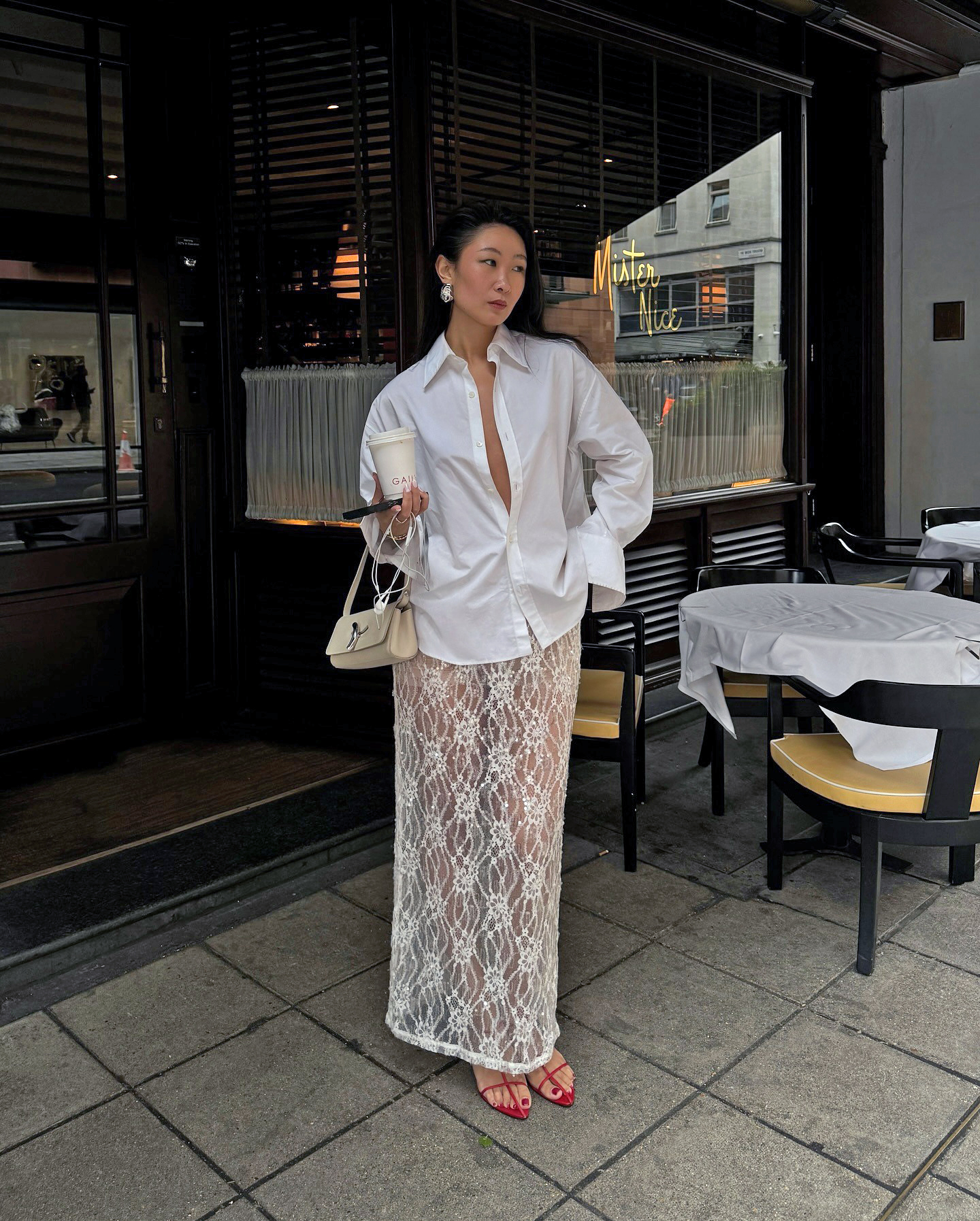 influencer Caroline Lin poses outside Mister Nice in London wearing an unbuttoned white button-down shirt with wide cuffs, small cream shoulder bag, white sheer lace maxi skirt and strappy red heeled sandals