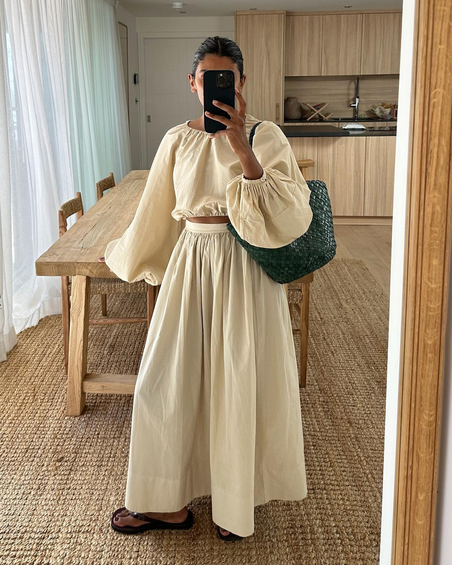 British style influencer Monikh Dale poses for a mirror selfie in the living room of her London home wearing a beige linen smocked neck cropped top with balloon sleeves and a matching linen skirt, green Dragon Diffusion tote bag, and black flat flip-flop sandals
