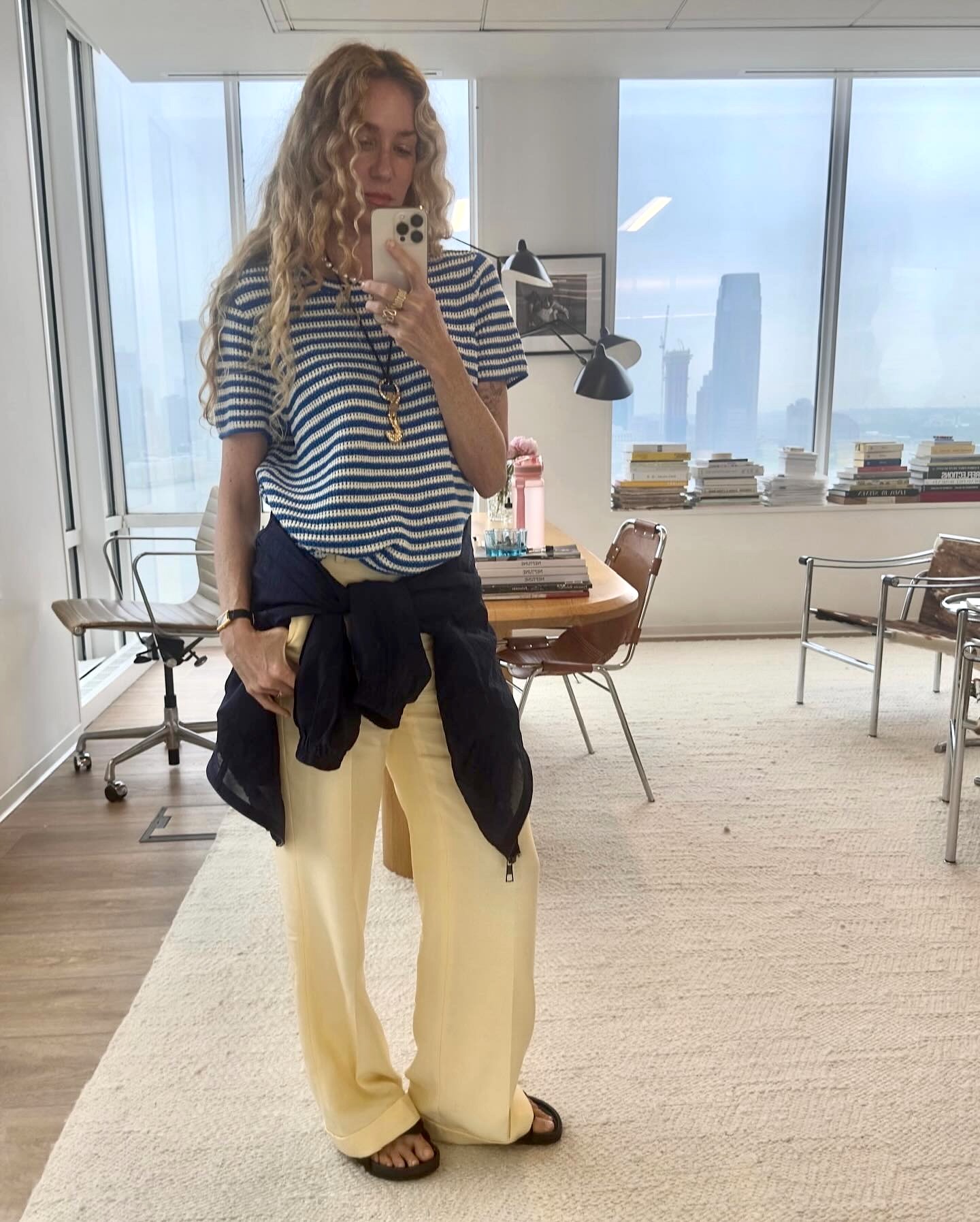 Olympia Gayot, creative director and head of design for J.Crew, poses for a mirror selfie in her office wearing a knit striped shirt, a long pendant necklace, a shirt tied around her waist, butter-yellow linen pants, and flat sandals.