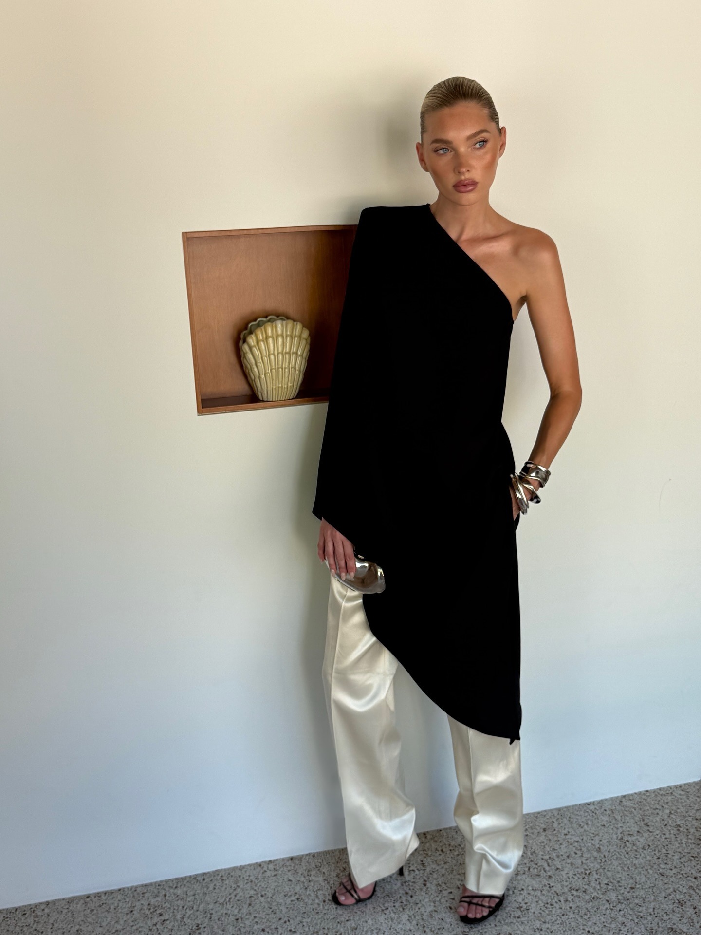 Elsa Hosk styled multiple bangles on her wrist with a black one-sleeved drape top over white satin trousers and black heeled sandals.