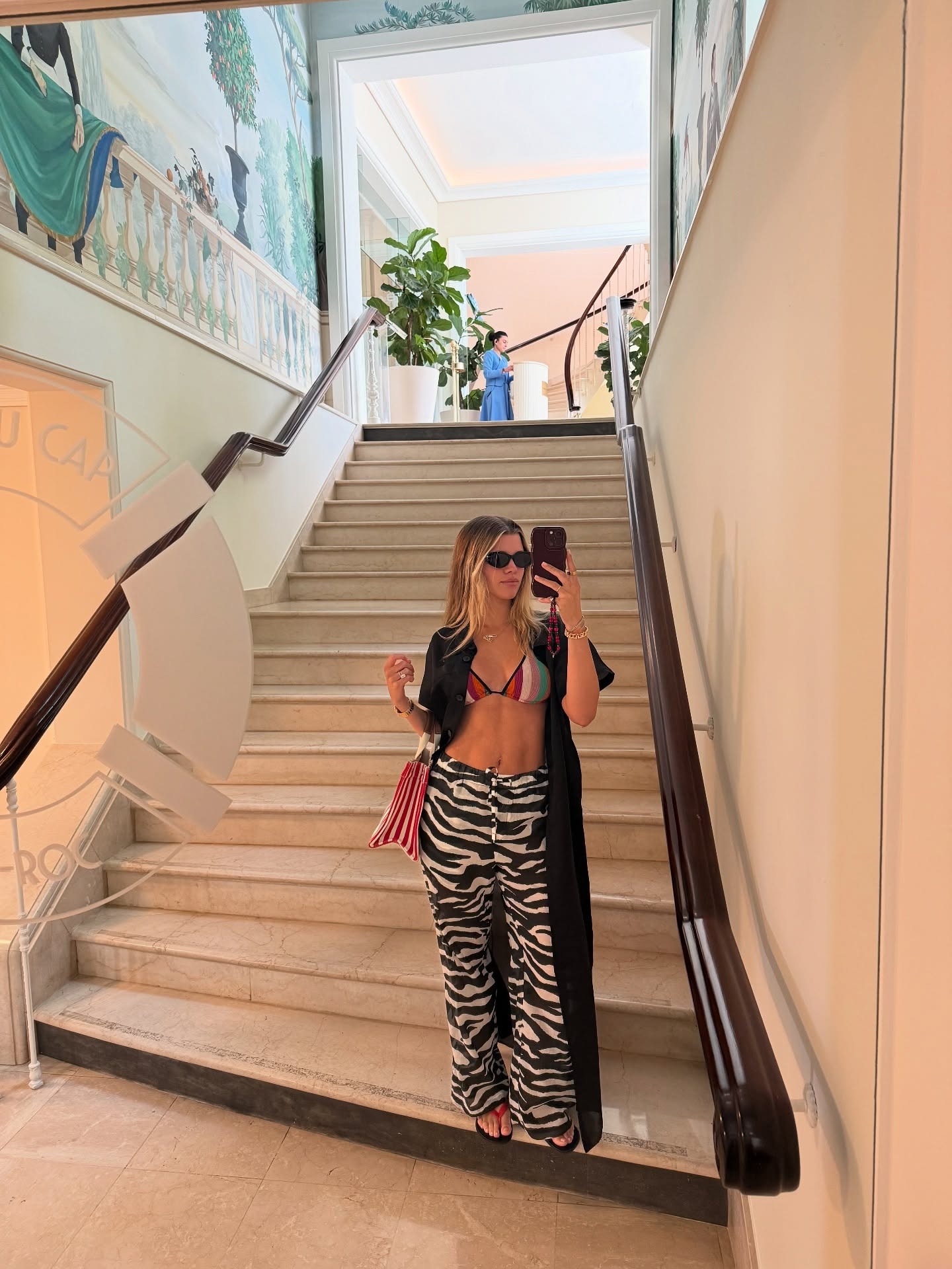 Sofia Richie Grainge stands at the bottom of a staircase taking a selfie in a mirror, wearing zebra print trousers with a multicoloured, striped bikini top and red flip-flops.