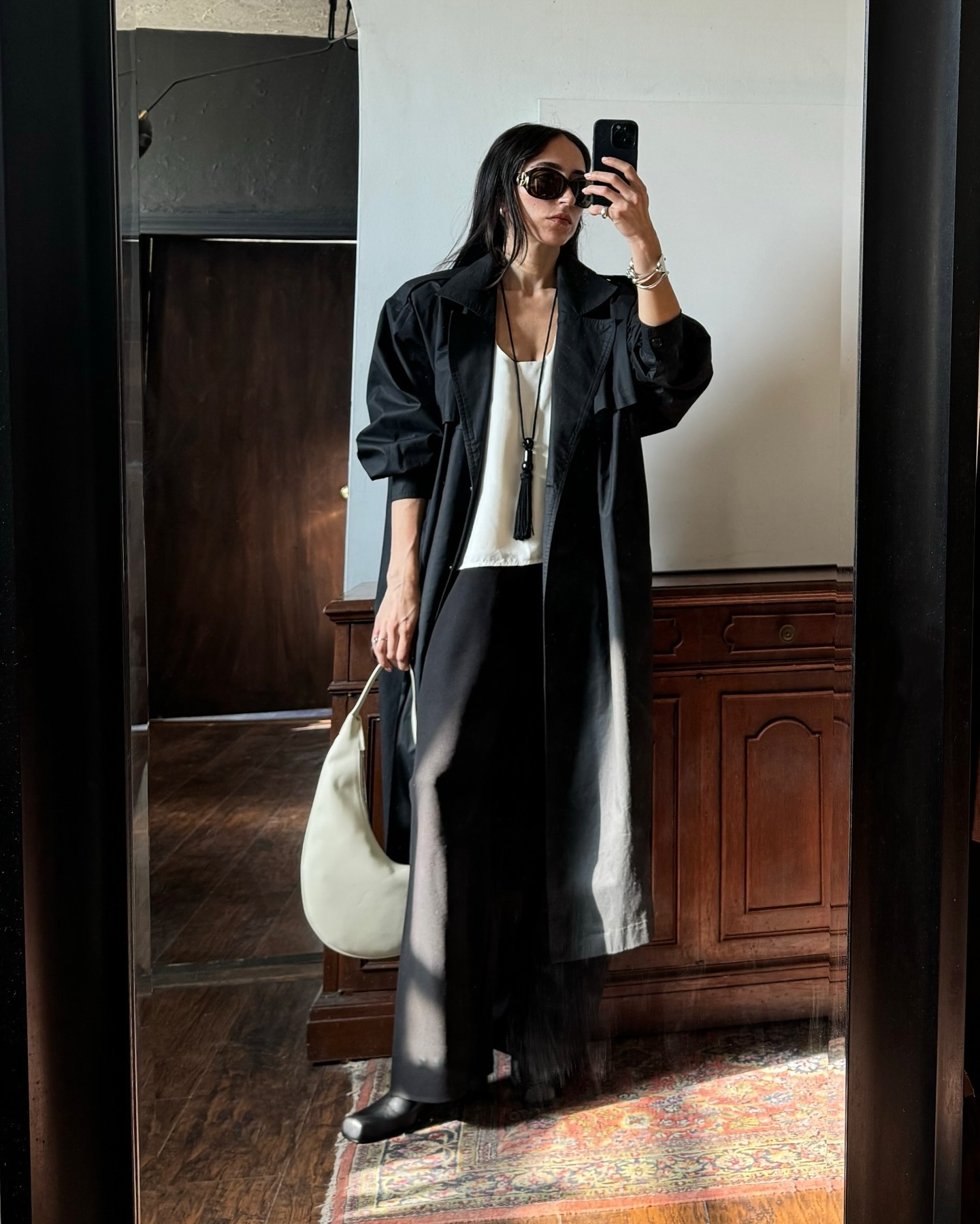 Alexis wears a black coat, white top, black pants, a black tassel necklace, and black sunglasses.