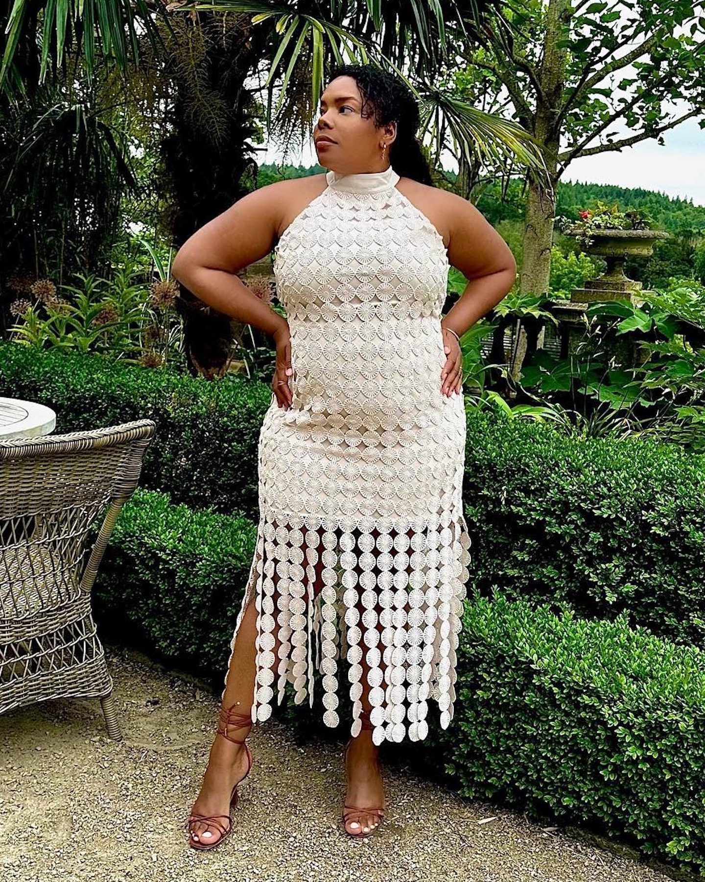 London based influencer Chloé Pierre poses outside the Beaverbrook Hotel & Spa in the UK wearing a white halter dress with a fringed skirt and neutral camel brown ankle strap strappy heeled sandals