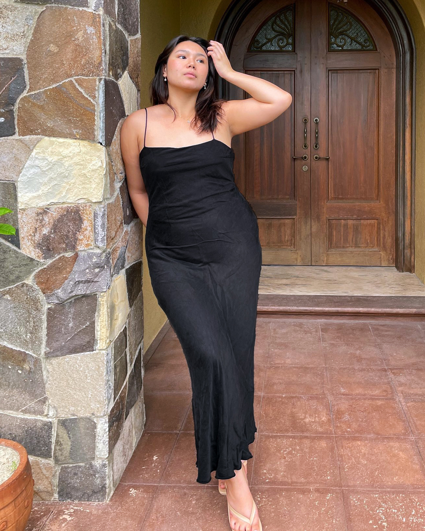 mid-size fashion influencer Marina Torres leans against a stone wall wearing a black cami maxi slip dress with neutral heeled flip-flop sandals
