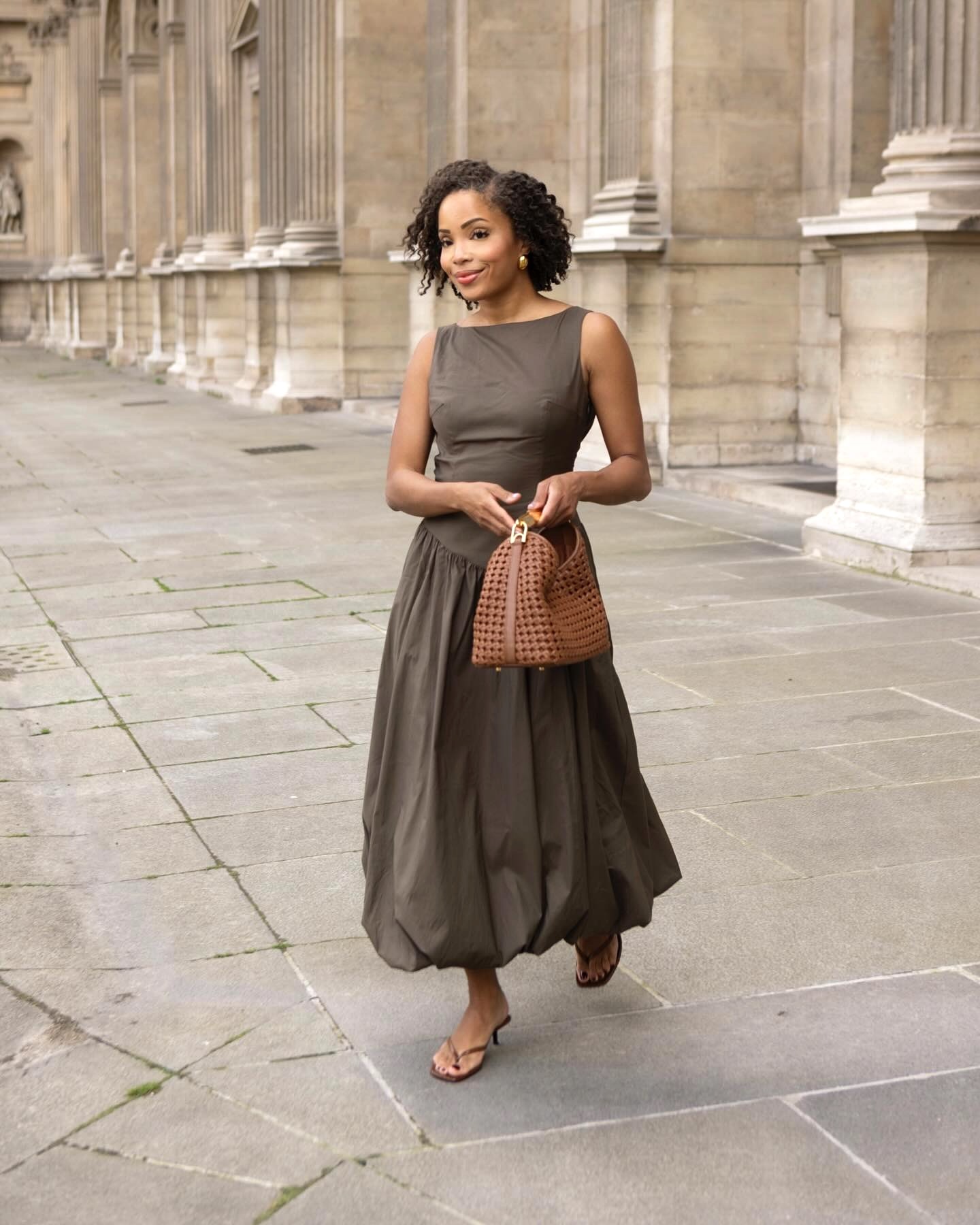 Paris influencer Ellie Delphine walking holding a camel bag, brown sleeveless maxi bubble-hem dress and neutral brown heeled flip-flop thong sandals