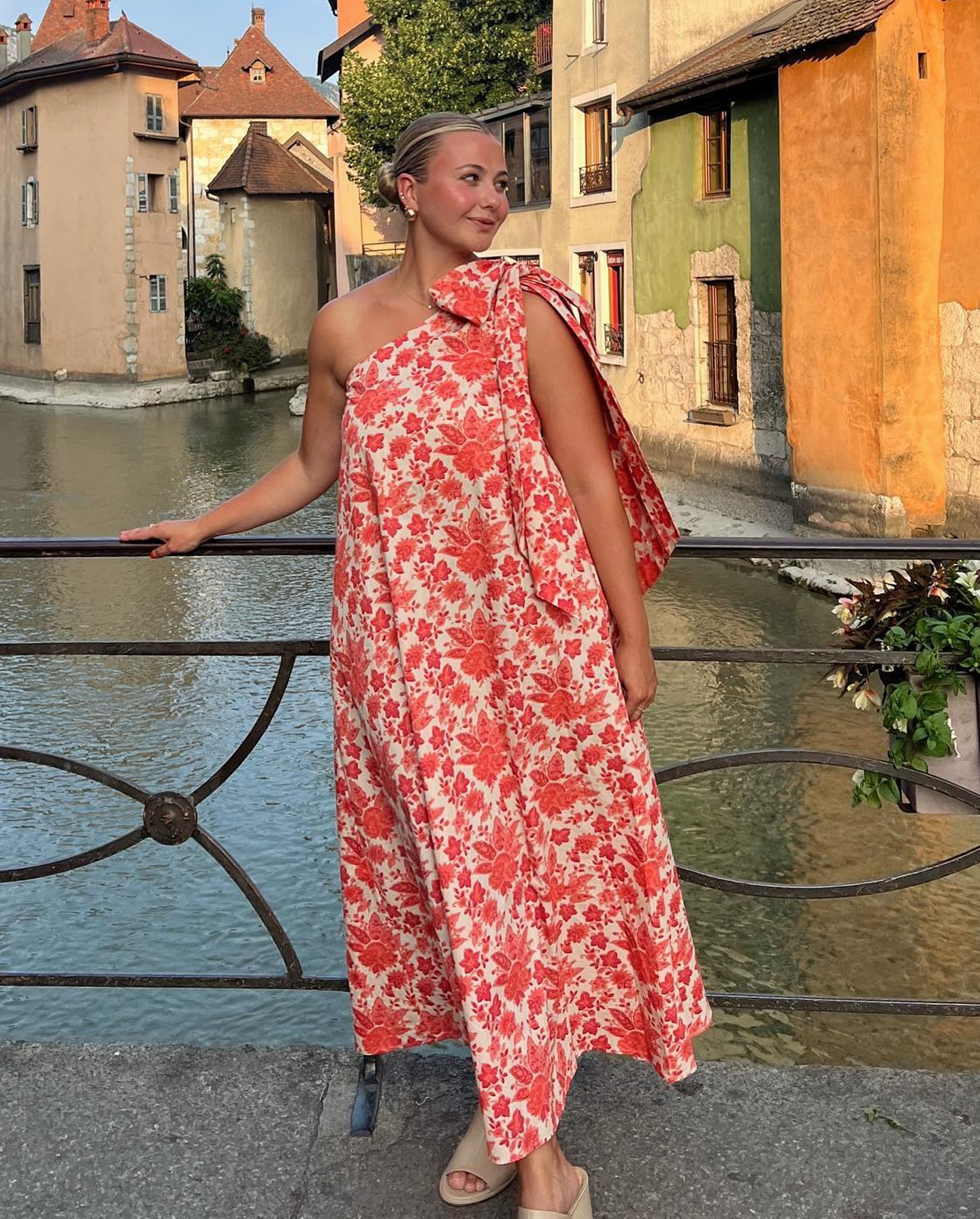 fashion influencer Olivia Marcus poses on a bridge in Lake Annecy, France wearing a bright floral-print one-shoulder maxi dress and neutral tan mule heels