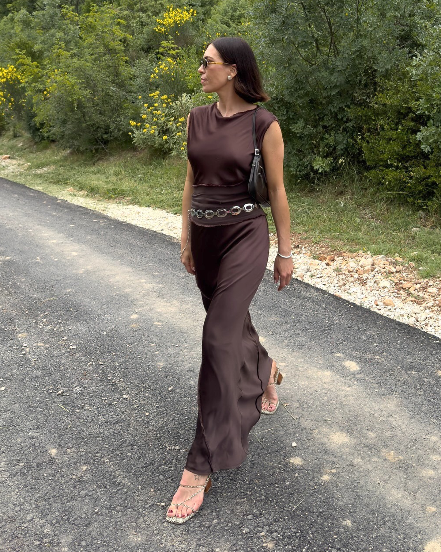 British influencer Jessica Skye wearing sunglasses, a small black shoulder bag, brown silky sleeveless dress, metal loop accent belt, and strappy neutral snakeskin heeled sandals