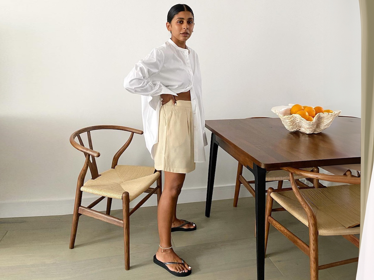 British influencer Monikh Dale poses in her chic London dining room with an abstract white bowl full of oranges on the table wearing a white oversize line button-down shirt, butter-yellow shorts, anklets and black flat flip-flop sandals