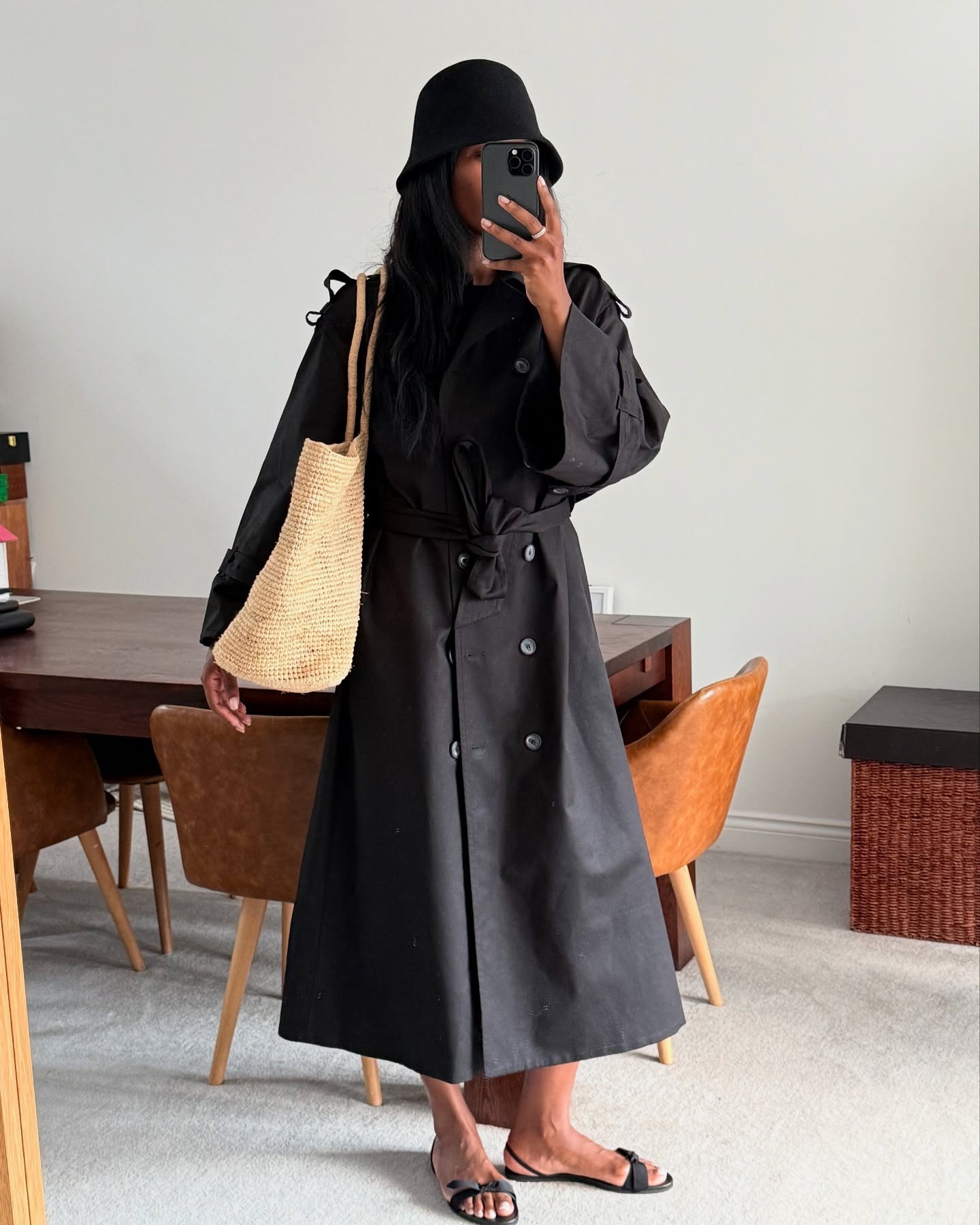 Marilyn wears a black hat, black trench coat, black flats, and a straw tote bag.