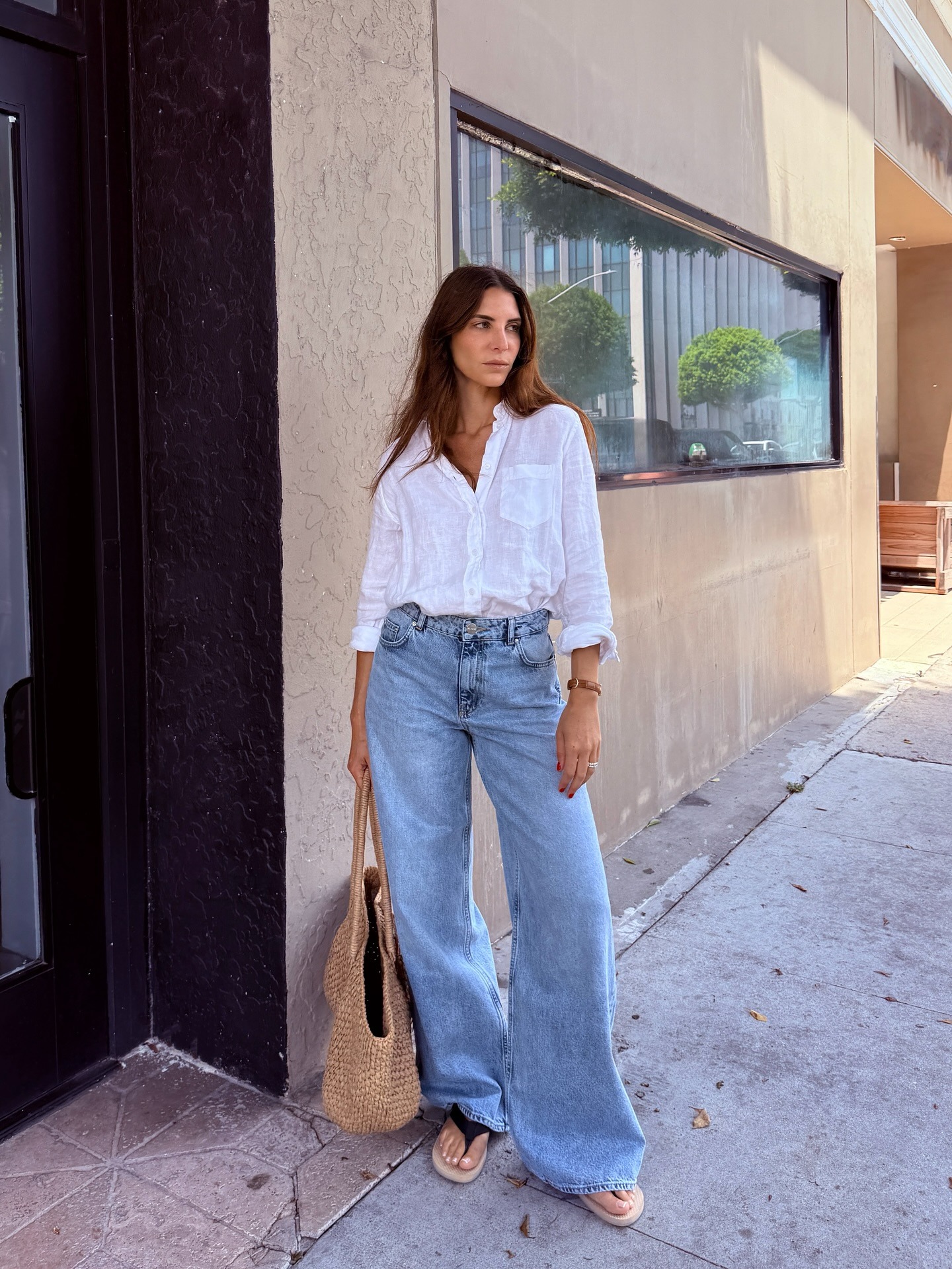 Jen wears a white button down, jeans, flip flops, and a straw tote bag.