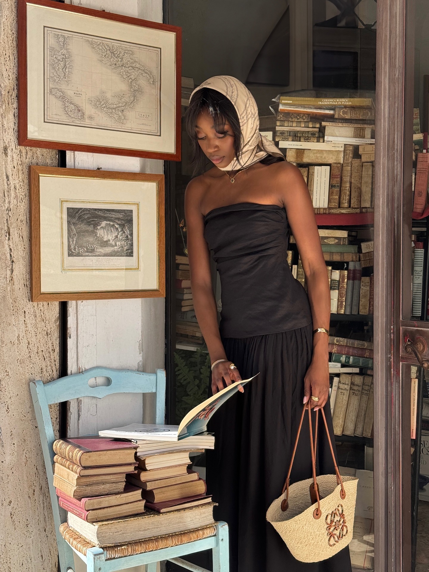 Emmanuelle wears a scarf around her hair, a black strapless dress, and a loewe straw tote.