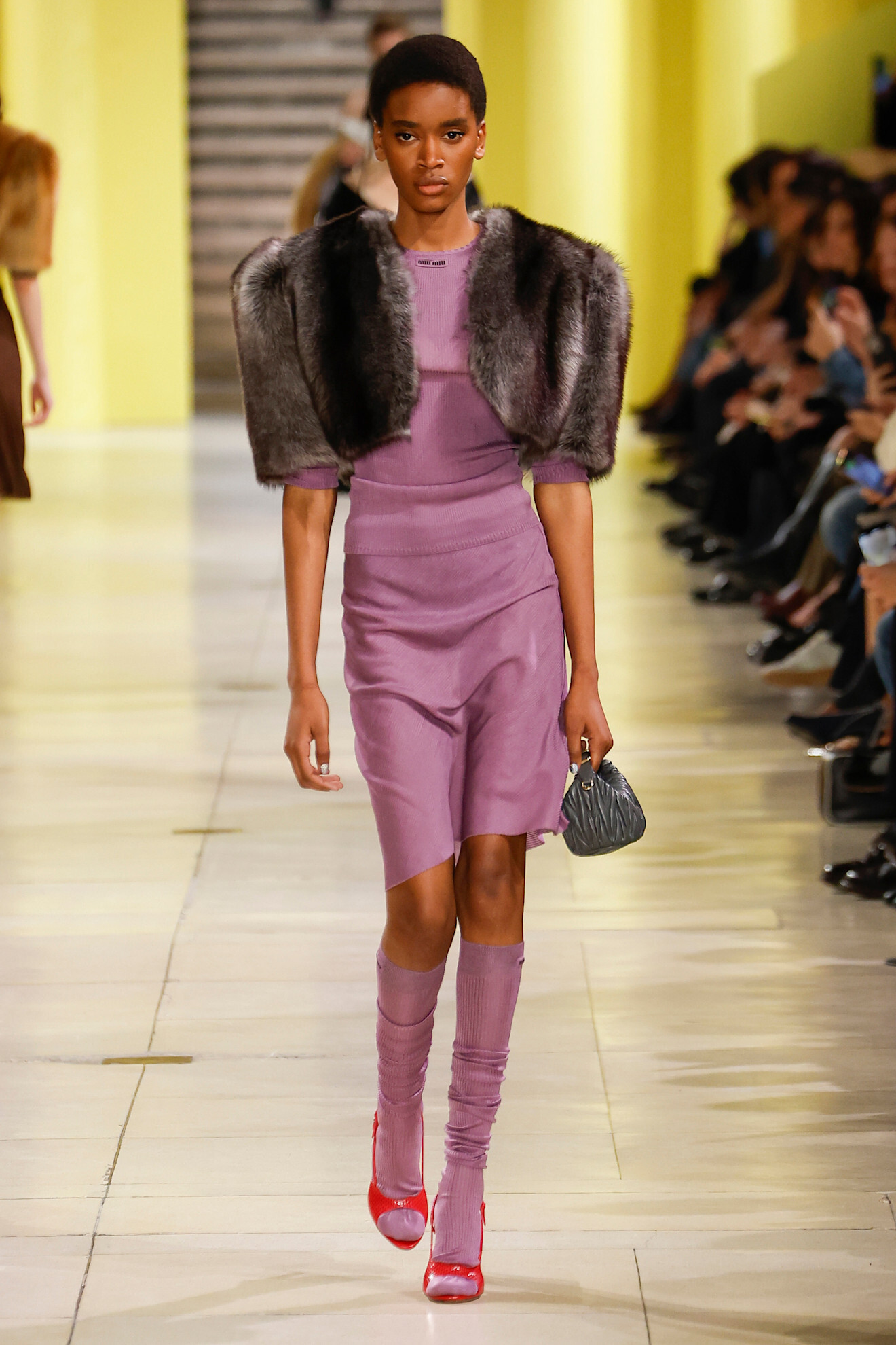 A model wearinga short fur jacket, pink dress, and sheer socks on the Miu Miu runway