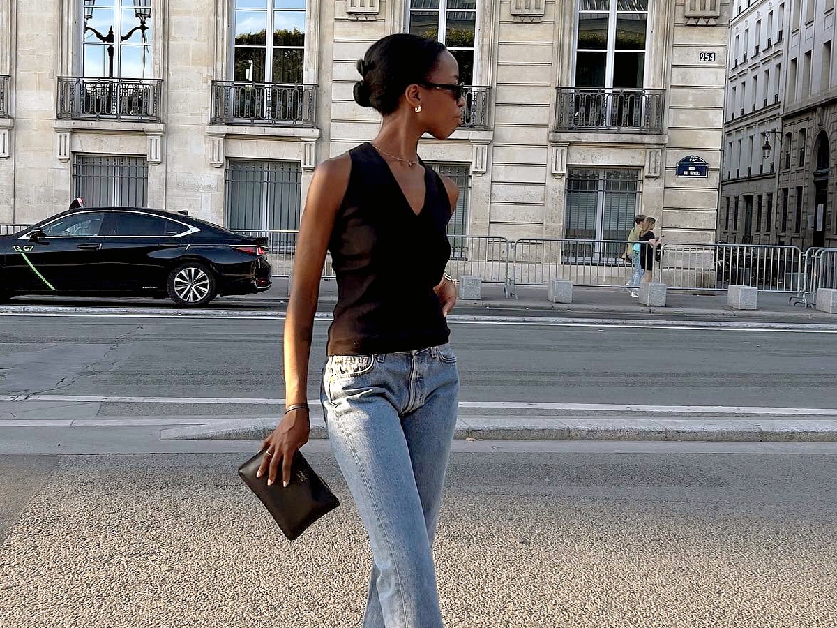 British influencer Sylvie Mus poses on a sidewalk in the 1st arrondissement in Paris wearing black sunglasses, gold hoop earrings, a v-neck black sleeveless top, black clutch, and straight-leg jeans