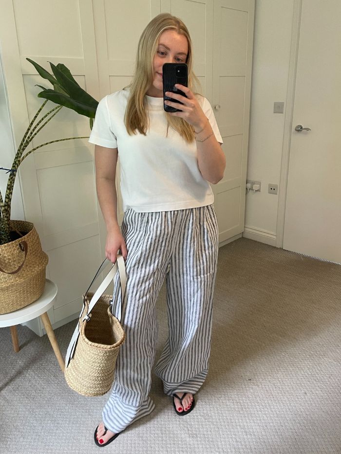 Woman wears White t-shirt, striped linen trousers, black sandals and basket bag
