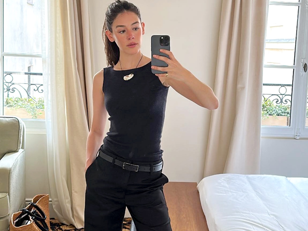 style influencer Ilana Torbiner poses for a mirror selfie in her hotel room at Château Voltaire in France wearing a black cord necklace with a silver bean pendant, a black boatneck tank top, black belt, and black pants