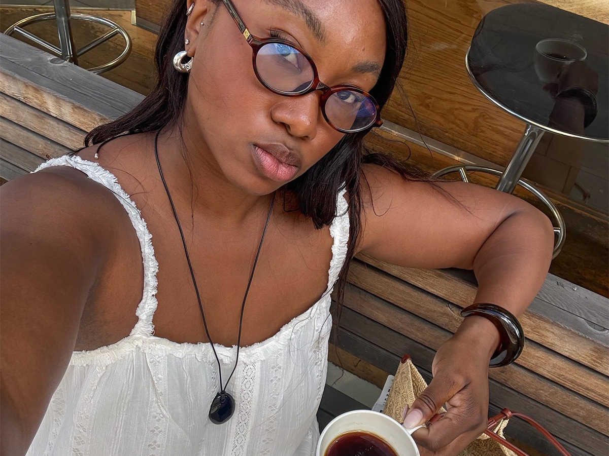 Influencer Nnenna wears a white lace dress and a long pendant necklace while holding a cup of coffee.
