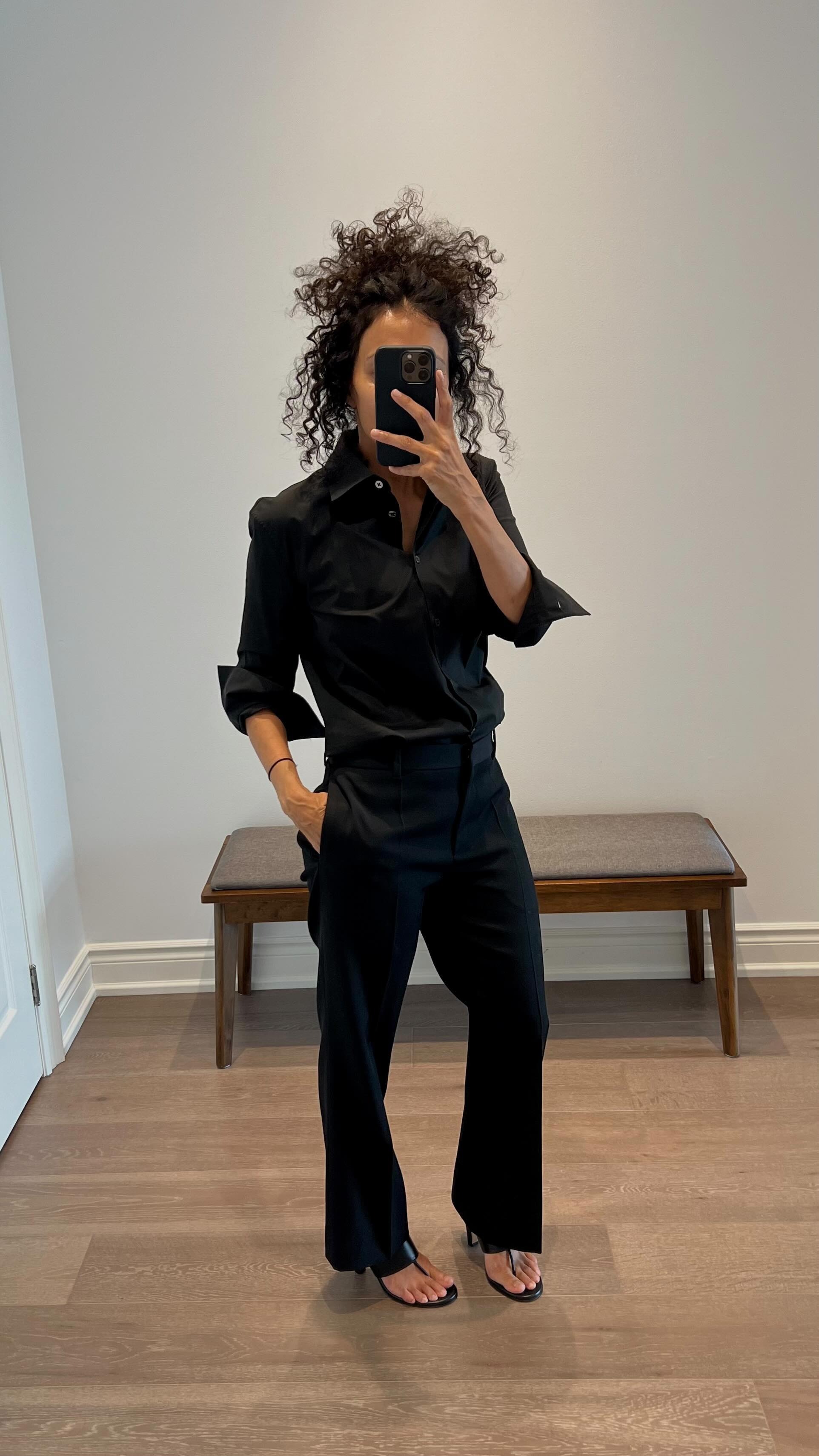 Neelam wears a black button-down top, black trousers, and black thong heeled mules.