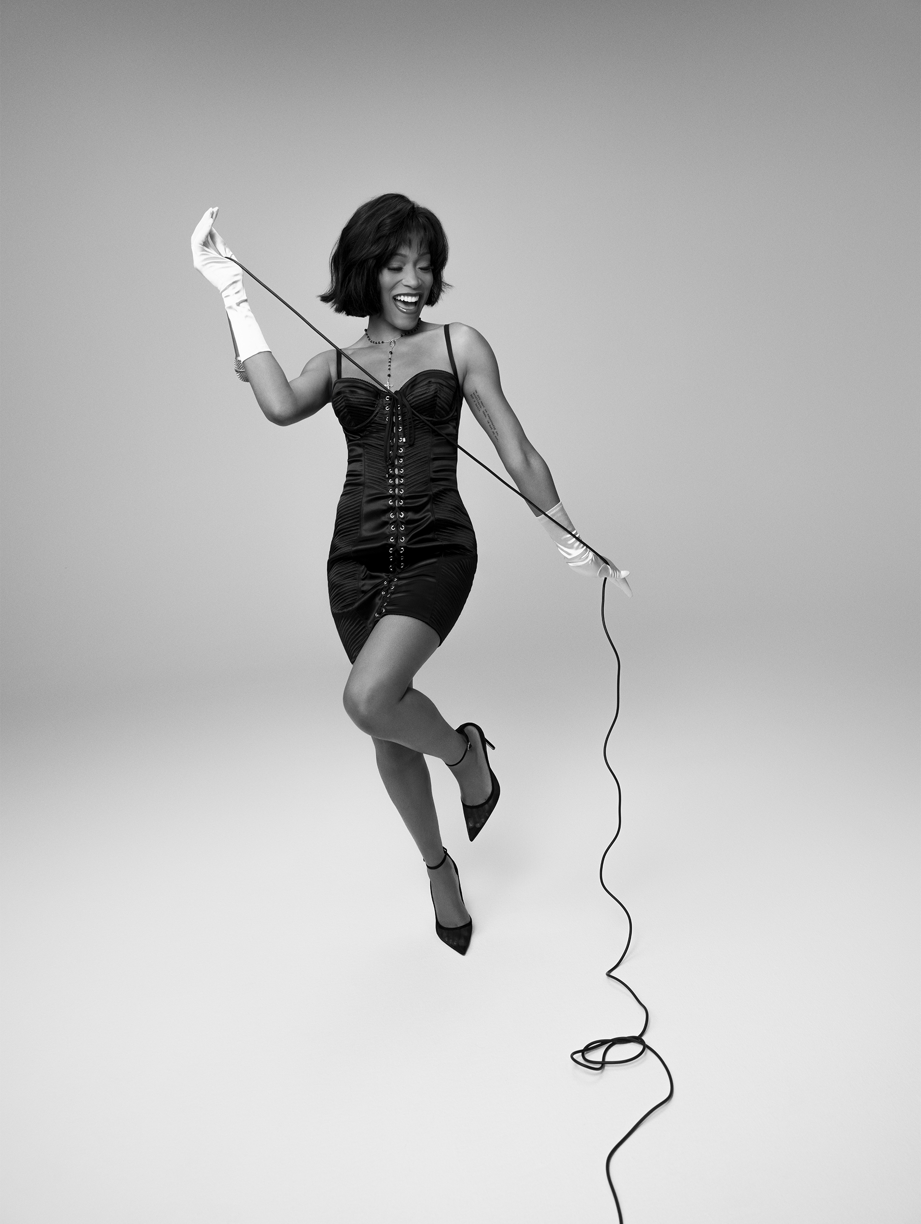 Keke Palmer references Whitney Houston for Who What Wear's July cover story. She is wearing a black bustier dress that laces up the front and green gloves. She is holding a microphone.