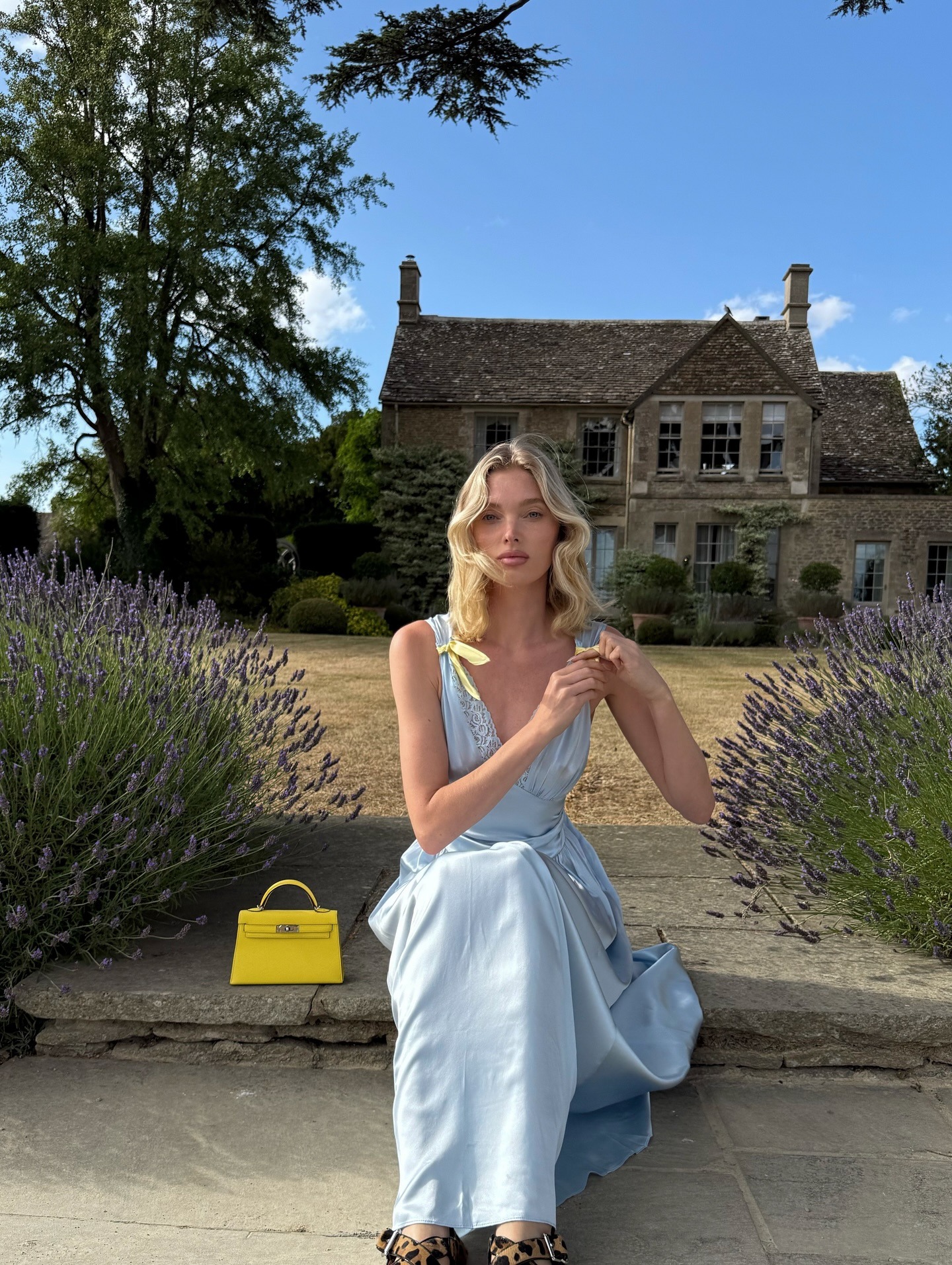 Elsa Hosk sits outside in a garden in front of an English countryside house wearing a blue satin maxi dress with yellow bows on the arms and a yellow Hermes bag.
