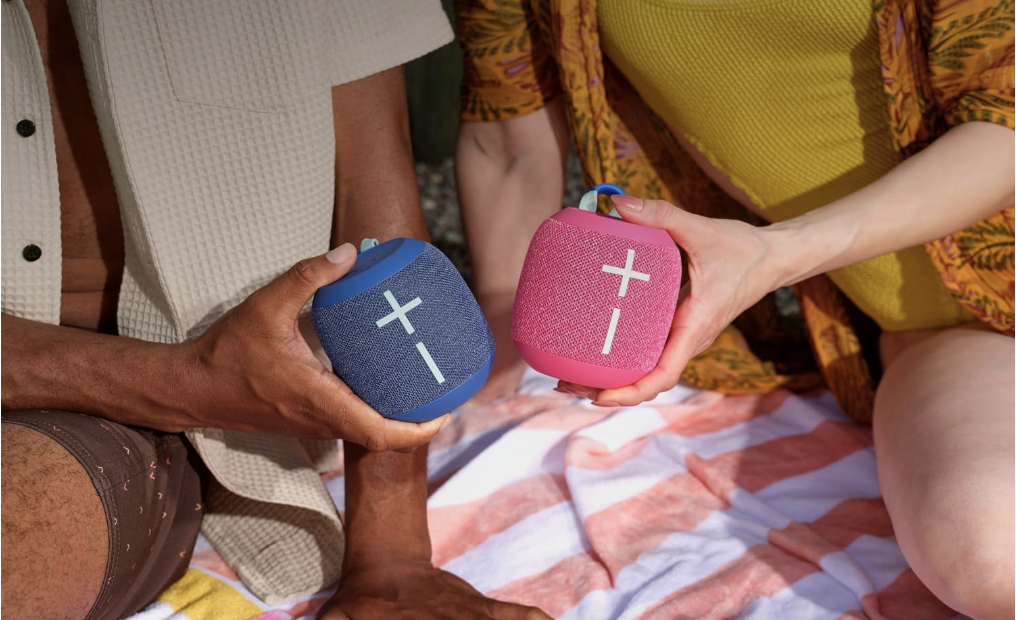 Two people holding UE WonderBoom 4 speakers, one blue and one pink.