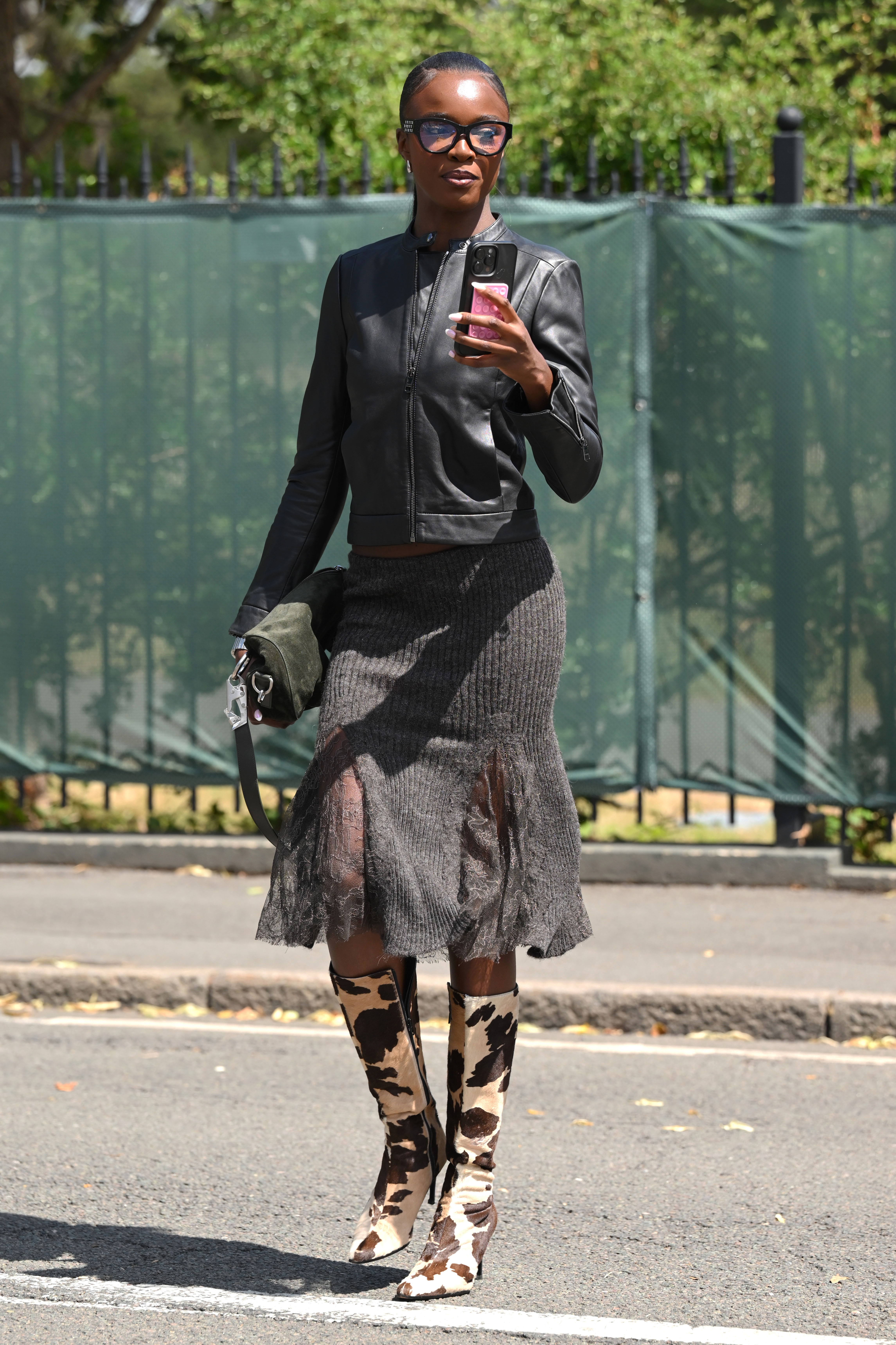 Leomie Anderson wearing a black leather jacket, a knit and lace grey skirt, black reading glasses, a green purse, and holding an iPhone in London